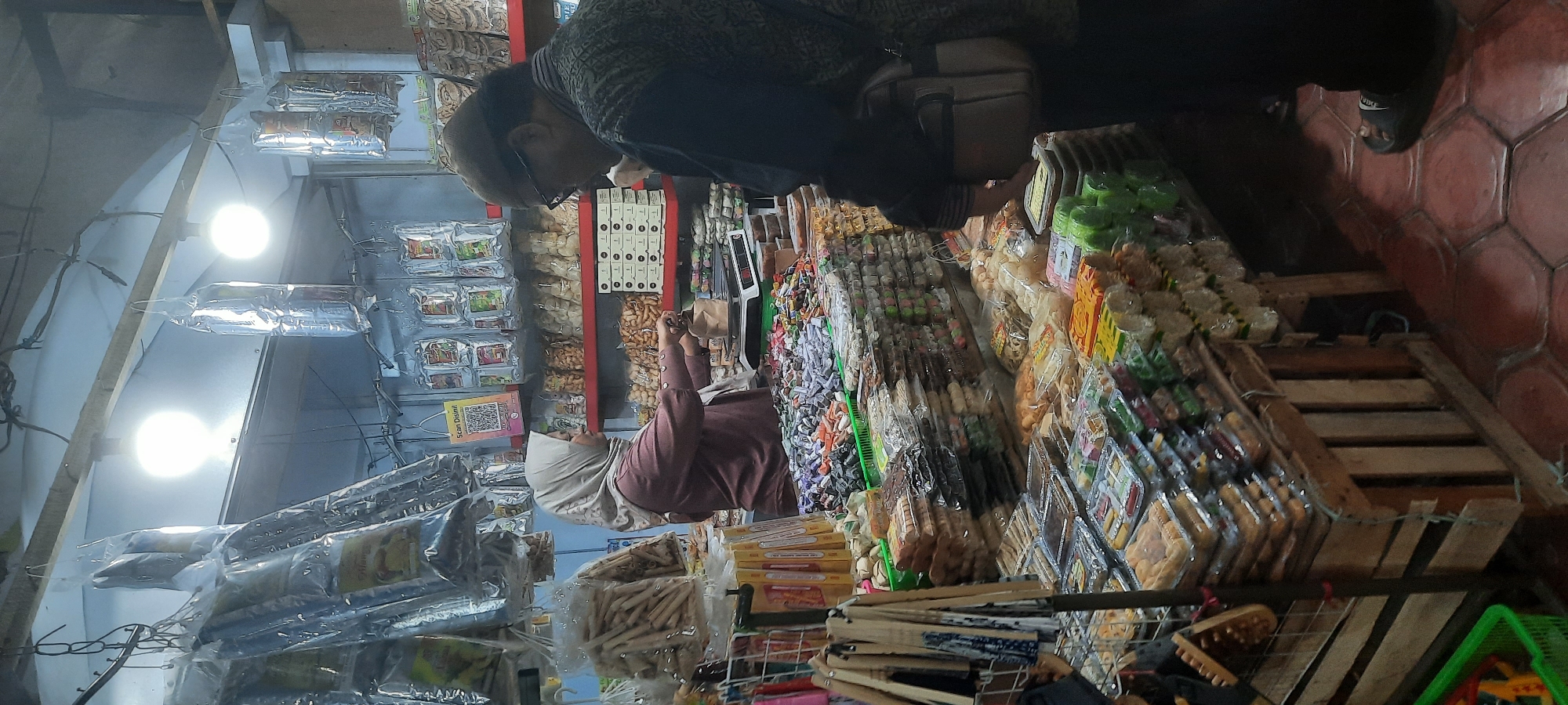 In the city of Blitar, Indonesia, a vendor wearing a light-colored hijab and a mauve shirt is operating a scale at a market stall. She is seen interacting with a customer, a man with glasses and a dark patterned shirt, who is facing away from the camera and appears to be observing the transaction. The stall is filled with various food items, including an abundance of individually wrapped candies and snacks displayed in clear plastic packaging. Some items are also hanging in larger plastic bags from the stall's overhead structure. The overall environment suggests a busy market setting, with a focus on confectionery and packaged snacks. The flooring consists of hexagonal tiles.