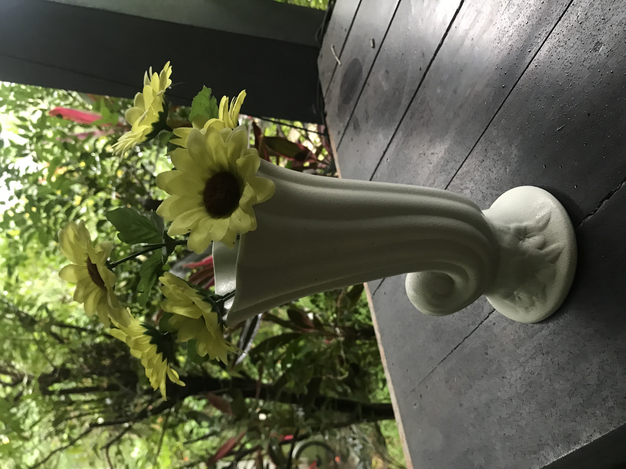 A white, fluted vase sits on a dark wooden surface outdoors. The vase contains yellow artificial sunflowers with green leaves. Behind the vase is a blurred background of green trees and foliage. The location appears to be a patio or porch with a garden view. The surface is dark, possibly stained wood. The image was taken during daylight hours.