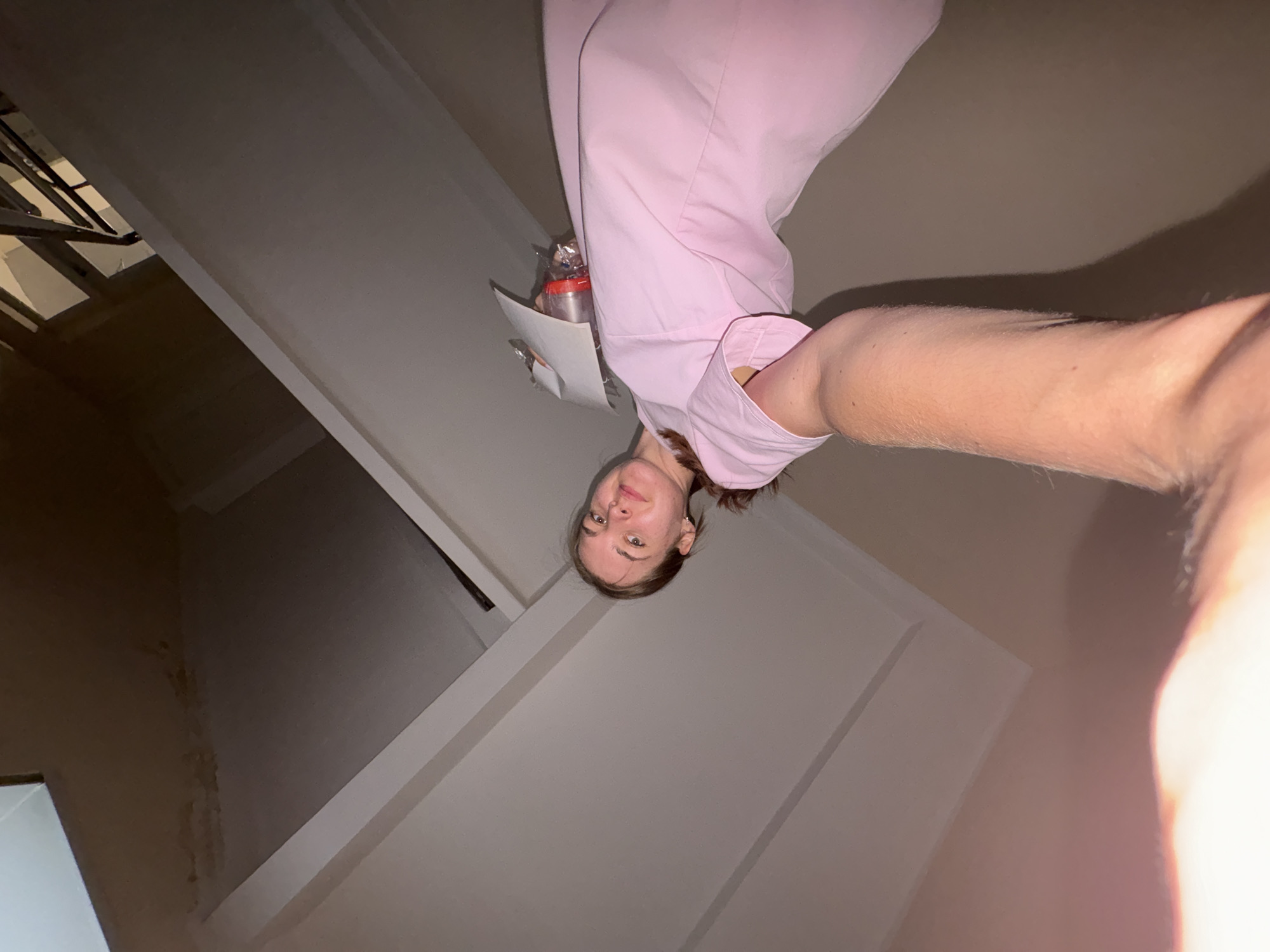 A selfie taken in what appears to be a stairwell. A woman holds the camera, with her arm visible in the foreground. She is wearing a pink top and holding papers and a small package. The stairwell has white walls. In the background, there is a glimpse of a metal railing and stairs. The photo's perspective is tilted. This photograph was taken indoors in Perm, Russia.