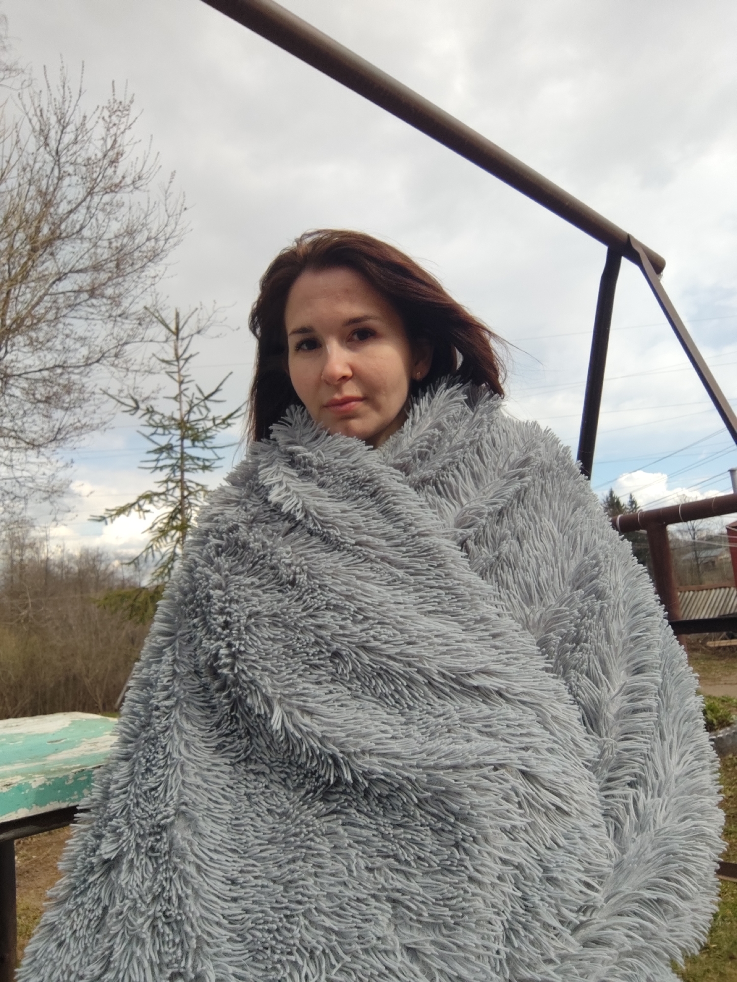 A woman is featured in a close-up outdoor shot, looking directly at the camera with a calm, neutral expression. She appears to be in her late 20s or early 30s, with long, dark brown hair that is slightly windblown. She is largely covered by a thick, fluffy grey blanket or wrap, which has a shaggy texture.
The scene is set outdoors, likely in a rural or semi-rural area consistent with Фоки, Russia. The sky is overcast with patches of brighter clouds, suggesting daytime, possibly during early spring or late autumn, given the bare branches of a large deciduous tree visible on the left side of the background. Below this tree, a smaller evergreen tree stands. On the far left, a weathered light green wooden surface, possibly a table or bench, is partially visible.
In the upper right portion of the image, a dark metal structure, possibly part of a swing set or an outdoor frame, cuts across the frame. A horizontal bar runs diagonally from the top left towards the right, and a supporting diagonal bar is also present. Faint utility lines can be seen in the background on the right, and distant buildings or structures are also vaguely discernible. The ground in the background appears to be dry grass or dirt. There is no visible text in the image.