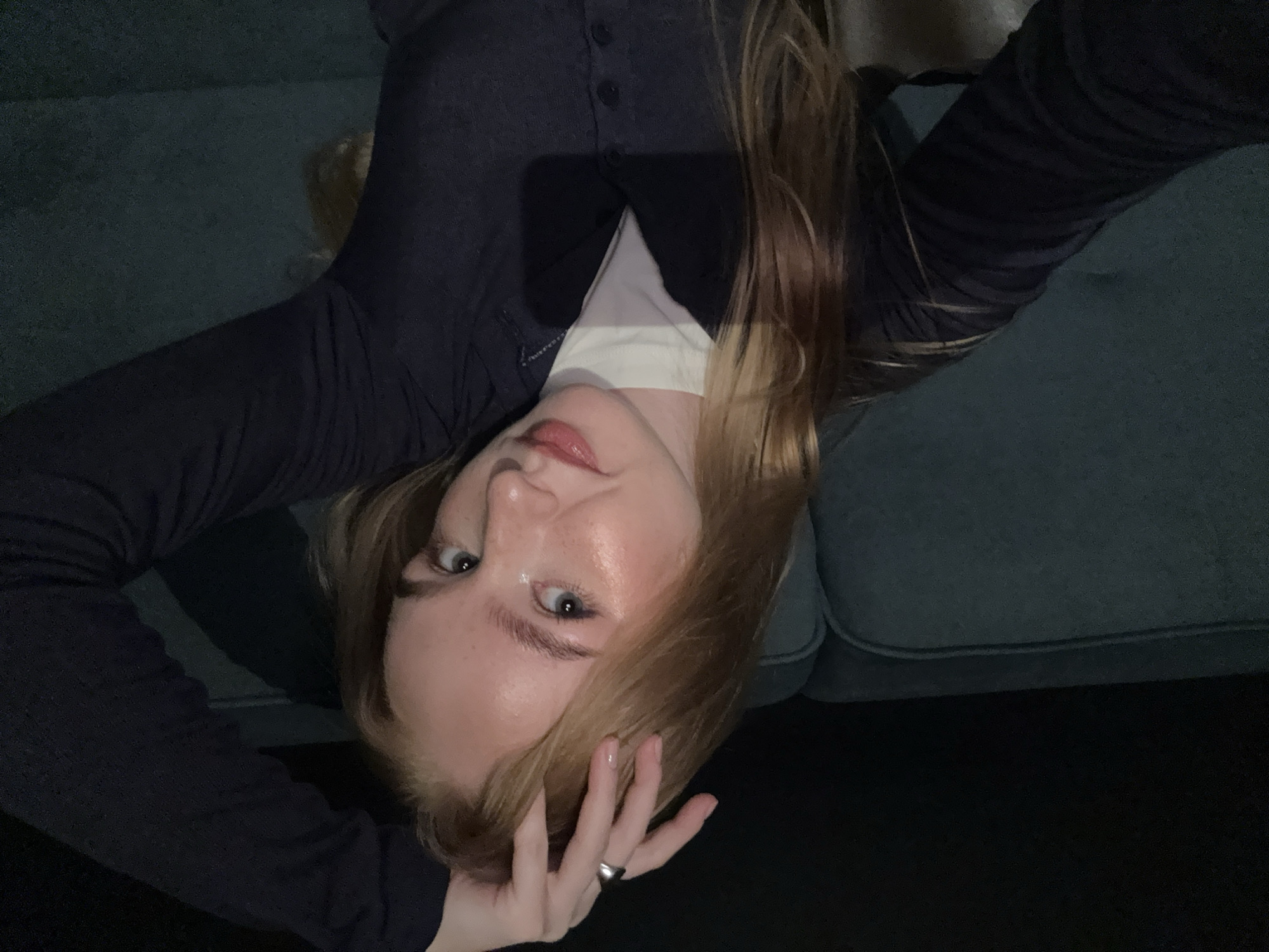 A young woman with blonde hair and blue eyes is posing for a selfie, her right hand on her head. She is wearing a dark blue cardigan over a white t-shirt and a silver ring on her right index finger. She is sitting on a dark teal couch, possibly in a home or an apartment, as indicated by the couch and the dark background. The lighting is dim, suggesting it is nighttime or a dark indoor setting. There is no information available to place this image in the city of Chaikovsky, Russia.