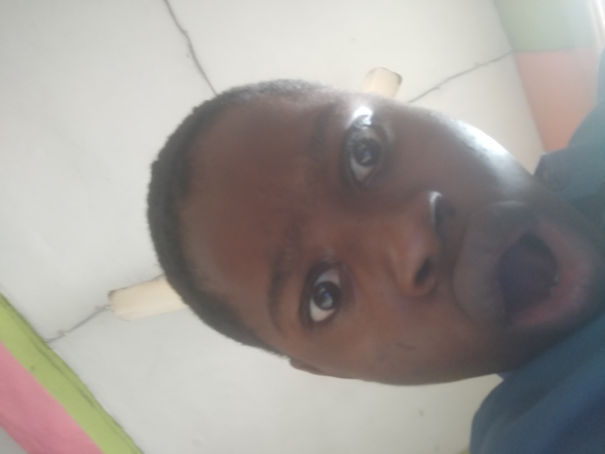 A close-up, low-angle selfie of a dark-skinned person with short hair. The person has their mouth open in a surprised or shouting expression. A ceiling with a light fixture and colorful stripes is visible in the background. The photo was taken in an indoor location, likely in Shangisha, Nigeria.