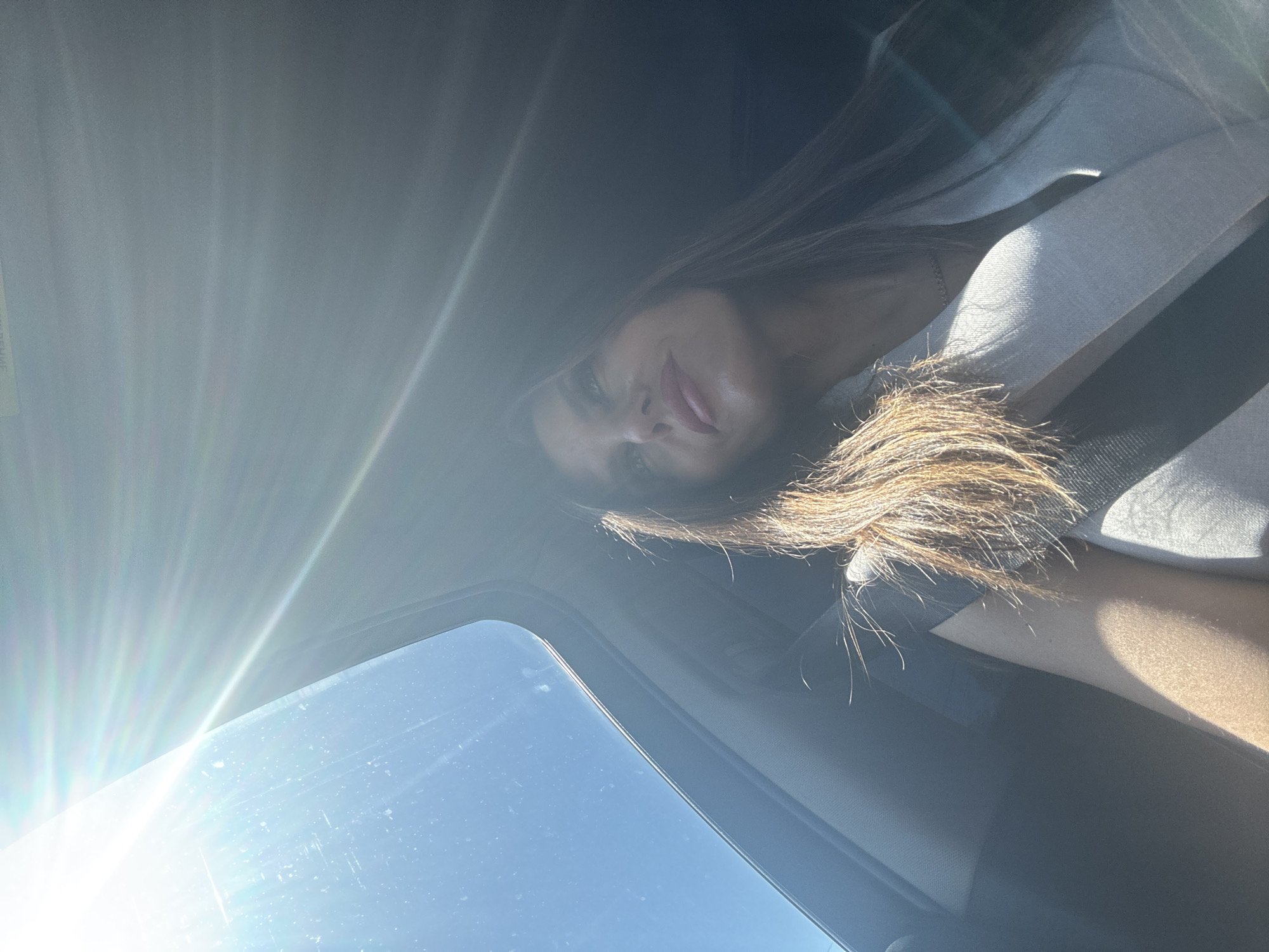 A woman is sitting in a car, possibly in Chaikovsky, Russia. The image is taken from a low angle, looking up towards the car's sunroof. The sun is bright, creating a glare. The woman is wearing a light-colored shirt with a black strap (likely a seatbelt). She has long brown hair. The sunroof shows a clear sky. The interior of the car is dark.
