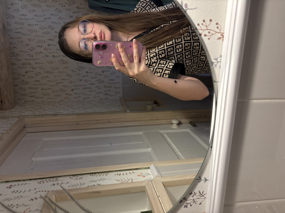 An indoor scene in Lezhnevo, Russia, depicting a female individual taking a mirror selfie. The subject is a woman with long, straight, light brown hair, wearing clear-framed eyeglasses and a short-sleeved top with a black "FF" monogram pattern on a beige background, featuring black cuffs. A delicate gold-colored bracelet with a small black four-leaf clover charm is visible on her right wrist. She holds a pink-cased iPhone with visible dual cameras with her right hand, positioned to capture her reflection. Her fingernails are painted a light pink. The reflection is captured in an oval mirror with a white decorative bottom frame and a light wood top-left trim, mounted above a white tiled surface. The background visible in the mirror features walls covered with wallpaper displaying a pattern of green stems and leaves with small red and orange accents on a light background. To the left of the subject's reflection, a light-colored panel door with a round doorknob and light wood trim is visible. A rough-hewn wooden beam runs along the ceiling line in the upper right portion of the reflection. A dark garment is partially visible hanging on the far right.