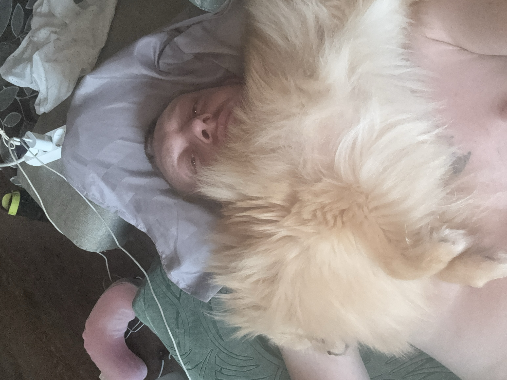 This is a close-up, overhead shot of a person lying down with a large, fluffy light-colored dog on top of them. The person's face is visible, and they appear to be looking slightly up and to the side. The dog's fur is a prominent feature, obscuring much of the person's body. The scene appears to be indoors, with bedding and possibly furniture visible in the background. There are some wires and what looks like a charger or electronic device near the edge of the frame. The overall mood seems relaxed and intimate, suggesting a moment of comfort between the person and their pet. The lighting suggests it is daytime.