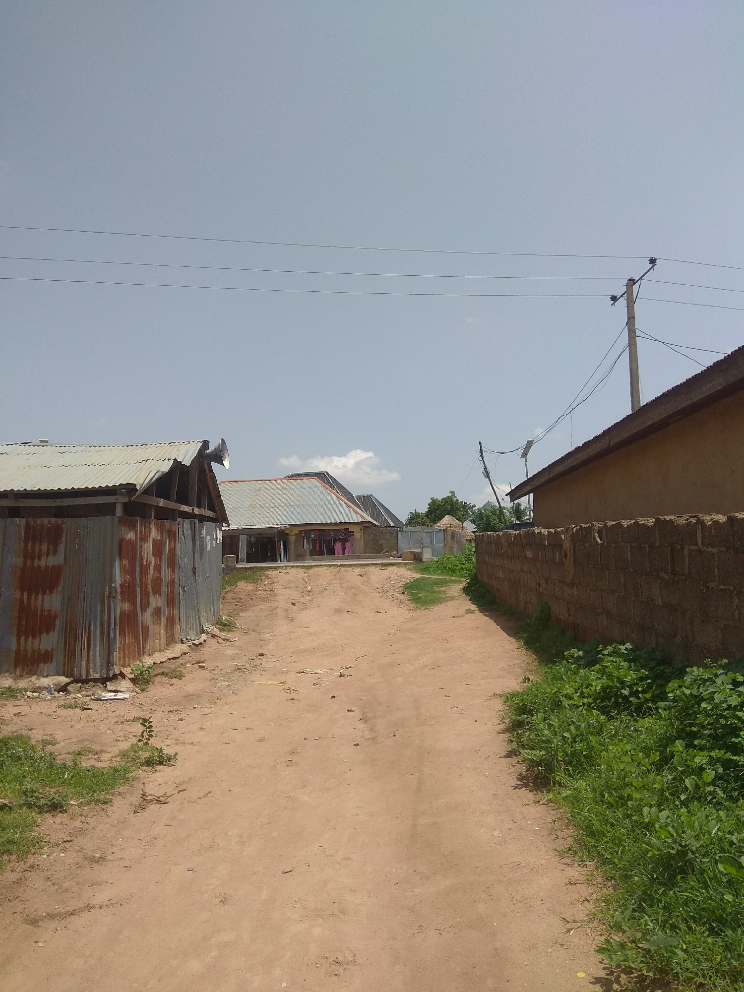 A dirt road leads through a residential area in Jalingo, Nigeria, under a clear, overcast sky. The road is a light brown color and appears to be unpaved. On the left side of the road, a corrugated metal structure with a rusty finish stands close to the foreground. It has a simple, angular roof. Further down the road on the left, another building with a corrugated metal roof is visible. Several items of clothing, including what appear to be pink shirts, are hanging on a line outside this building.
On the right side of the road, a wall constructed from rough, brown bricks runs parallel to the road. Beyond the wall, a building with a light brown, stucco-like exterior and a sloped, dark roof is situated. Overhead, power lines are strung between poles, with one pole visible on the right side of the image. Green vegetation, including grass and bushes, grows along the sides of the road and near the buildings. The overall impression is a simple, rural or semi-urban residential environment.