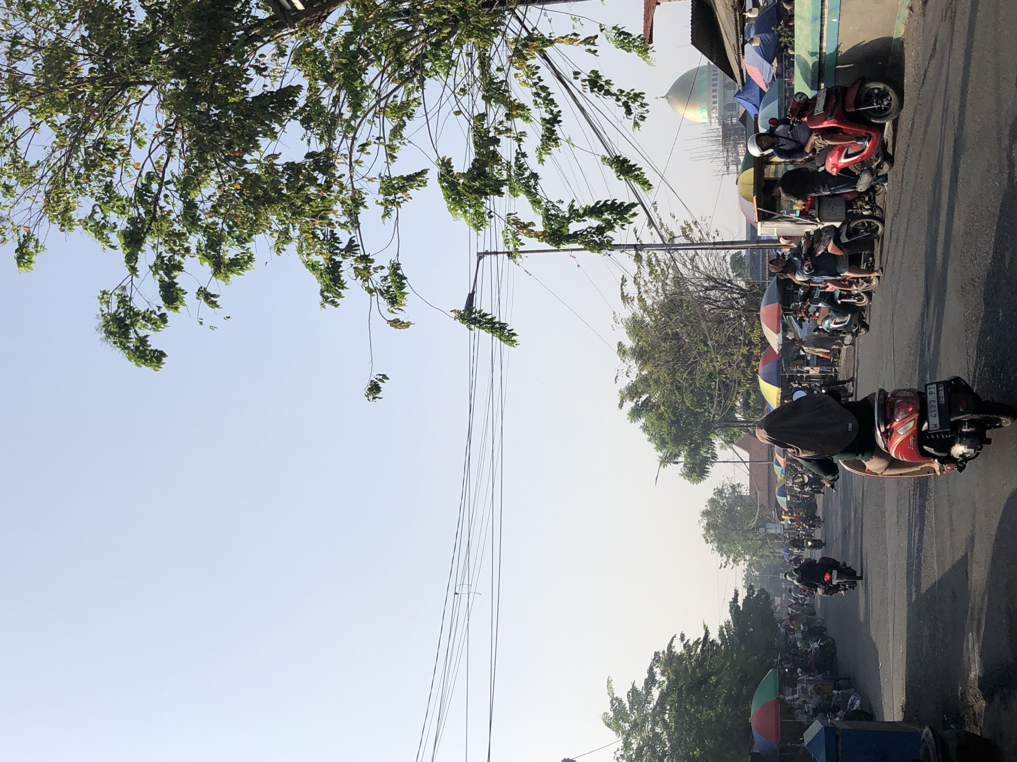 The image depicts a street scene in Balongdowo, Indonesia, under a clear, bright sky. A paved road is lined with a market on both sides, featuring numerous stalls covered by colorful umbrellas. People are seen riding motorcycles, walking, and standing around the market. A woman wearing a hijab rides a red motorcycle in the foreground, facing away from the camera. In the background, a golden dome structure, possibly a mosque, is visible above the market stalls and trees. Overhead, electrical wires crisscross the sky, supported by poles. The overall atmosphere suggests a bustling daytime activity in a local Indonesian setting.