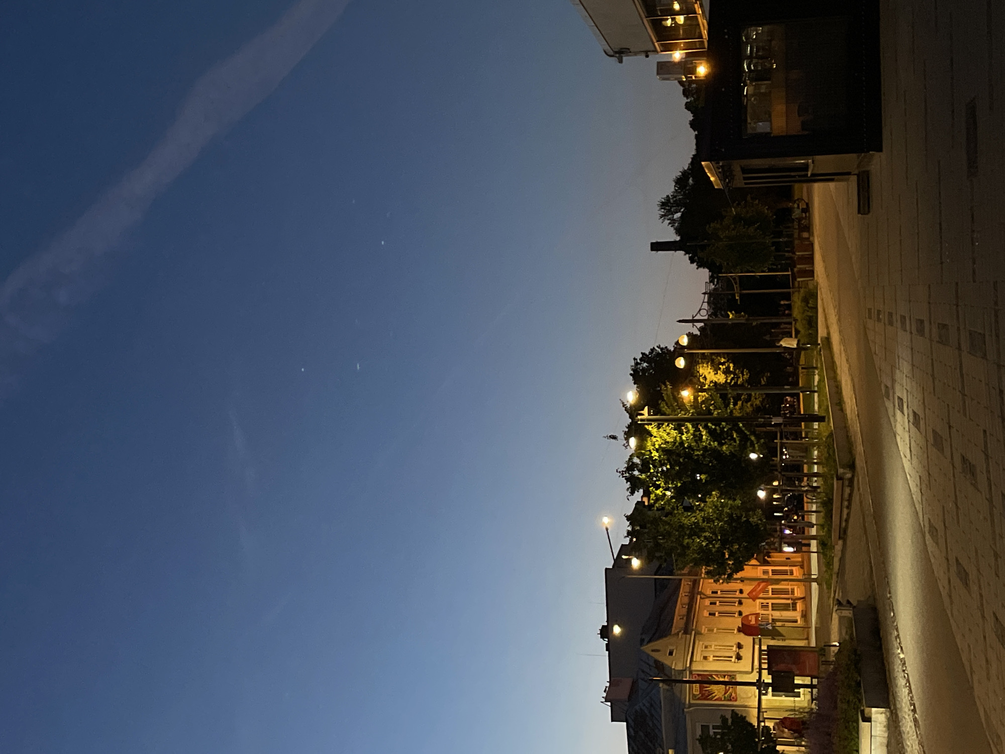 The image captures a street scene in Tallinn, Estonia during twilight. The sky is a deep blue, transitioning from lighter hues near the horizon. A faint contrail is visible in the upper right portion of the sky, and several small, bright dots, likely stars or distant lights, are scattered across the upper half of the frame.
In the middle ground, a row of buildings lines the left side of the street. The buildings are predominantly yellow and orange, with visible windows and architectural details. Streetlights with warm yellow bulbs illuminate the scene, casting light on trees and pathways. One building on the left has a prominent red banner hanging from its facade.
The foreground features a paved walkway with a brick-like pattern. The pathway is illuminated by streetlights and the ambient light from the buildings and sky. To the right, a dark, modern structure with glass windows stands prominently, possibly a shop or cafe. Beyond this, more trees and a glimpse of a darker building are visible.
A few indistinct figures are present in the mid-ground, walking or standing, but their details are not clear. The overall atmosphere is calm and serene, with the artificial lights creating a cozy ambiance against the darkening sky. The image appears to have been taken from a slightly elevated perspective, looking down the street.