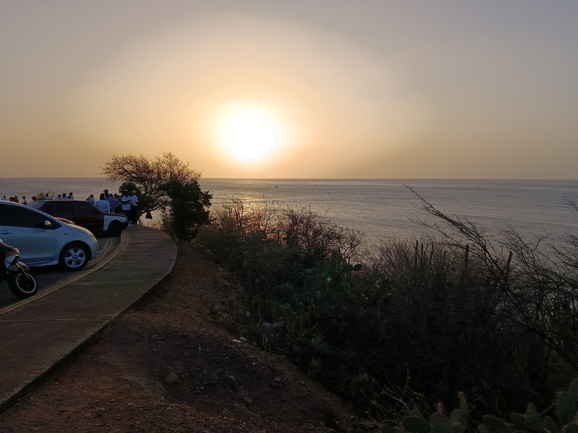 The image captures a sunset scene in Juan Griego, Venezuela, overlooking the ocean. The sun is positioned in the center of the sky, creating a bright glare reflecting on the water. A paved walkway lined with parked cars and a motorcycle is visible on the left side of the frame. A group of people are gathered near the cars, silhouetted against the setting sun. To the right of the walkway, there's a steep, rocky terrain covered with vegetation, including cacti and shrubs. The overall atmosphere is tranquil, with a warm, golden light illuminating the scene.