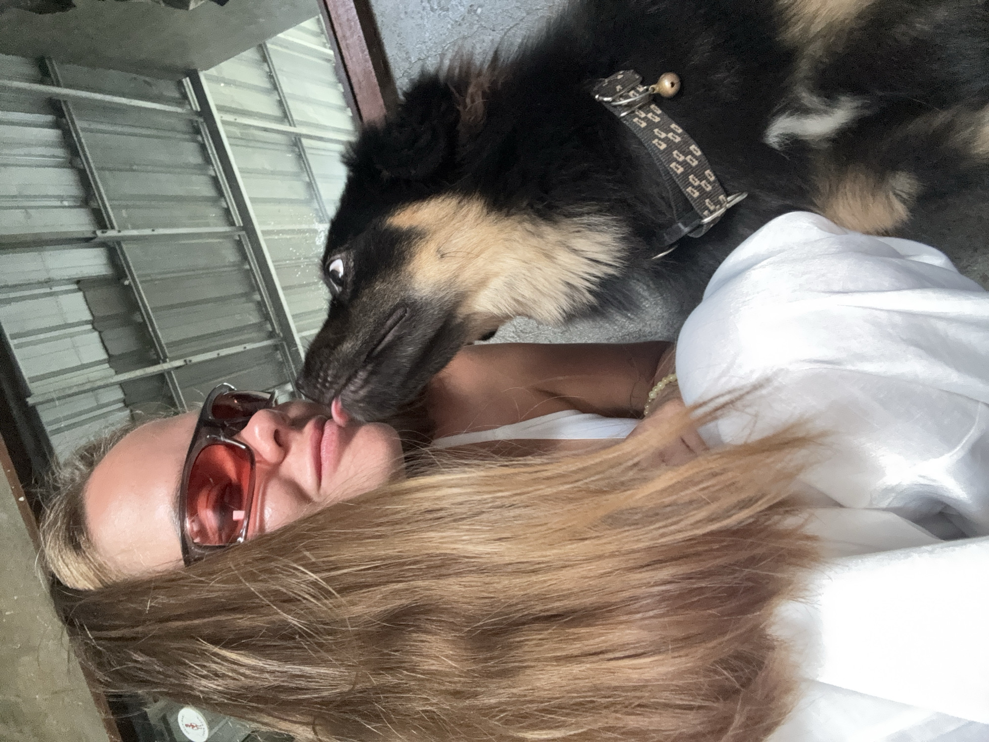 A fair-skinned woman with long blonde hair, wearing rose-tinted sunglasses, is lying down with a black and tan long-haired dog. The dog, wearing a patterned collar with a bell, is licking the woman's face. The background shows a corrugated metal structure with a frame. The location is North Kuta, Indonesia.