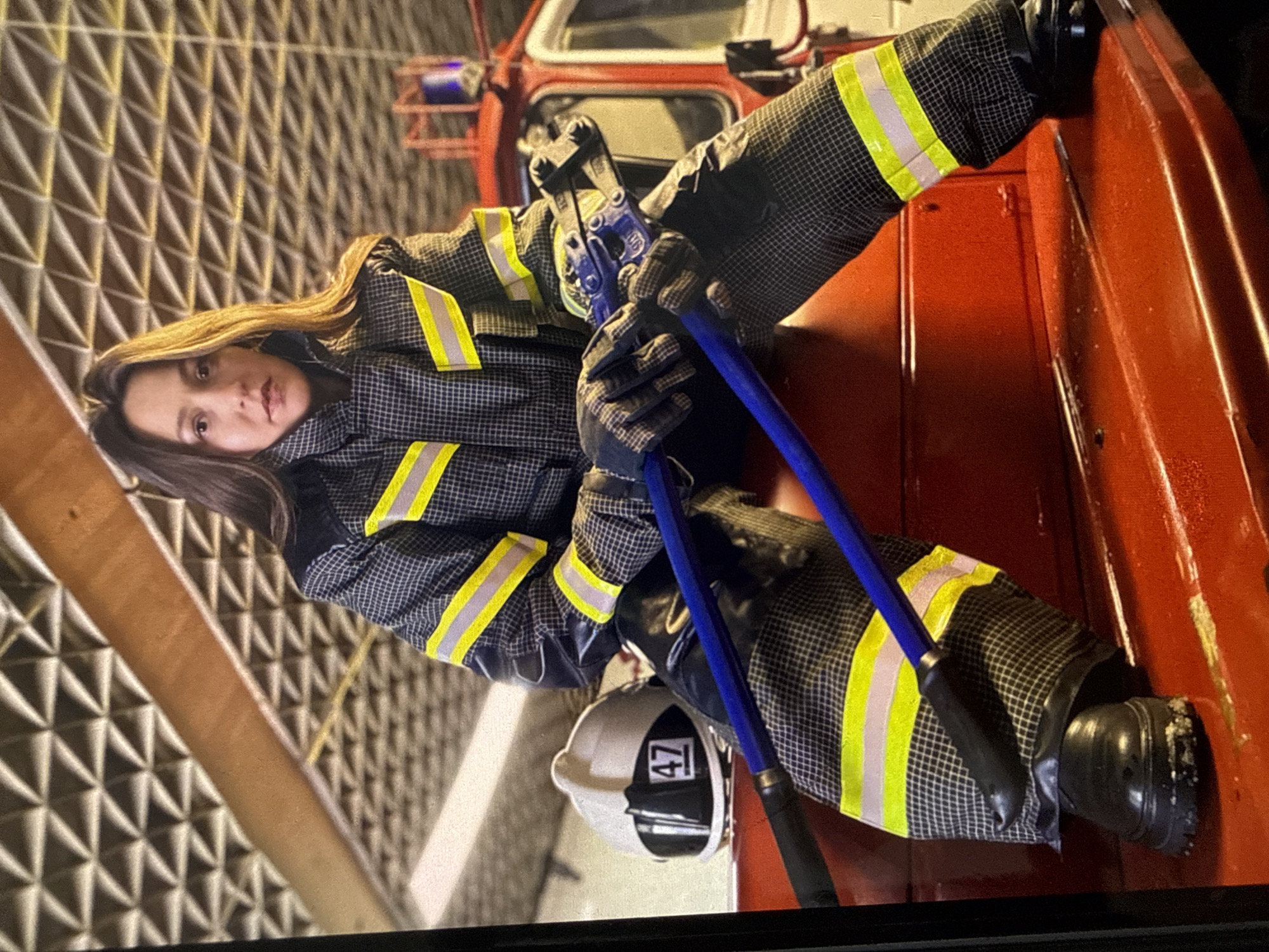 A woman wearing a dark grey and black patterned firefighter's uniform with yellow and silver reflective stripes is seated on the red metal surface of a fire truck. She is holding a pair of blue-handled bolt cutters. To her left, a white firefighter's helmet with a black stripe and the number "47" in a white box is visible. The background features a red fire truck with a blue light on top and a textured ceiling. The woman has long brown hair and is looking towards the camera with a neutral expression. The scene appears to be indoors, possibly in a fire station. There is no indication of the location being specifically in the city of Chaikovsky, Russia.
