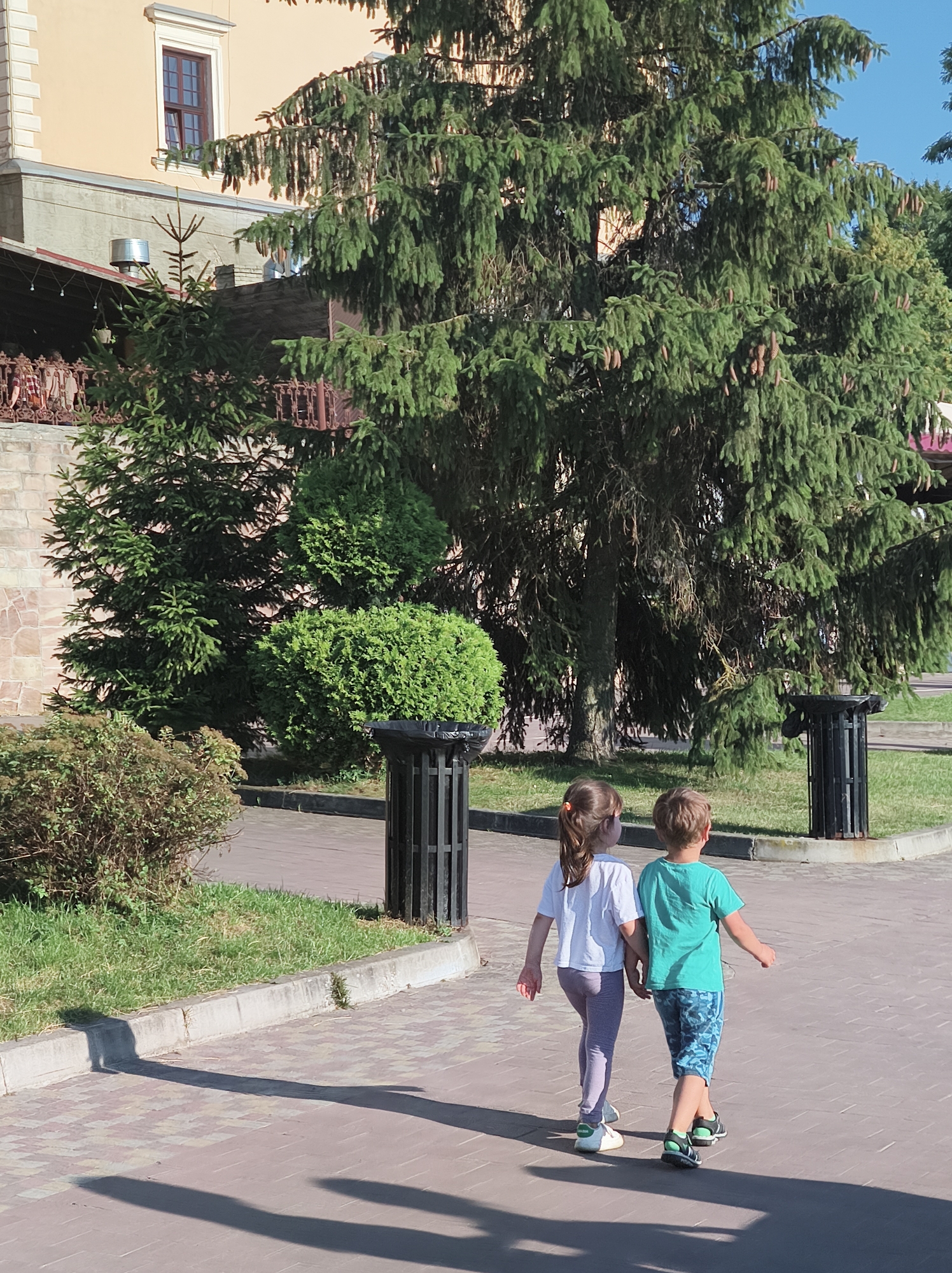 This is a medium shot, outdoor, eye-level, natural light photograph of two young children, a girl and a boy, walking hand-in-hand on a paved walkway. The girl, on the left, has long brown hair in a ponytail and is wearing a white t-shirt and light purple leggings. The boy, on the right, has short blond hair and is wearing a teal t-shirt and blue patterned shorts. They are walking away from the camera.
The background features a large evergreen tree, smaller manicured bushes, and a stone building with an ornate window. The walkway is paved with reddish-brown bricks. To the right of the children are two black trash receptacles. Green grass is visible beyond the walkway. The scene appears to be within the Ternopil National Economic University campus in Ternopil, Ukraine, potentially near the main building or one of the adjoining green spaces. The lighting suggests it is daytime, with strong shadows cast on the walkway, indicating a sunny day. The overall atmosphere is peaceful and casual.