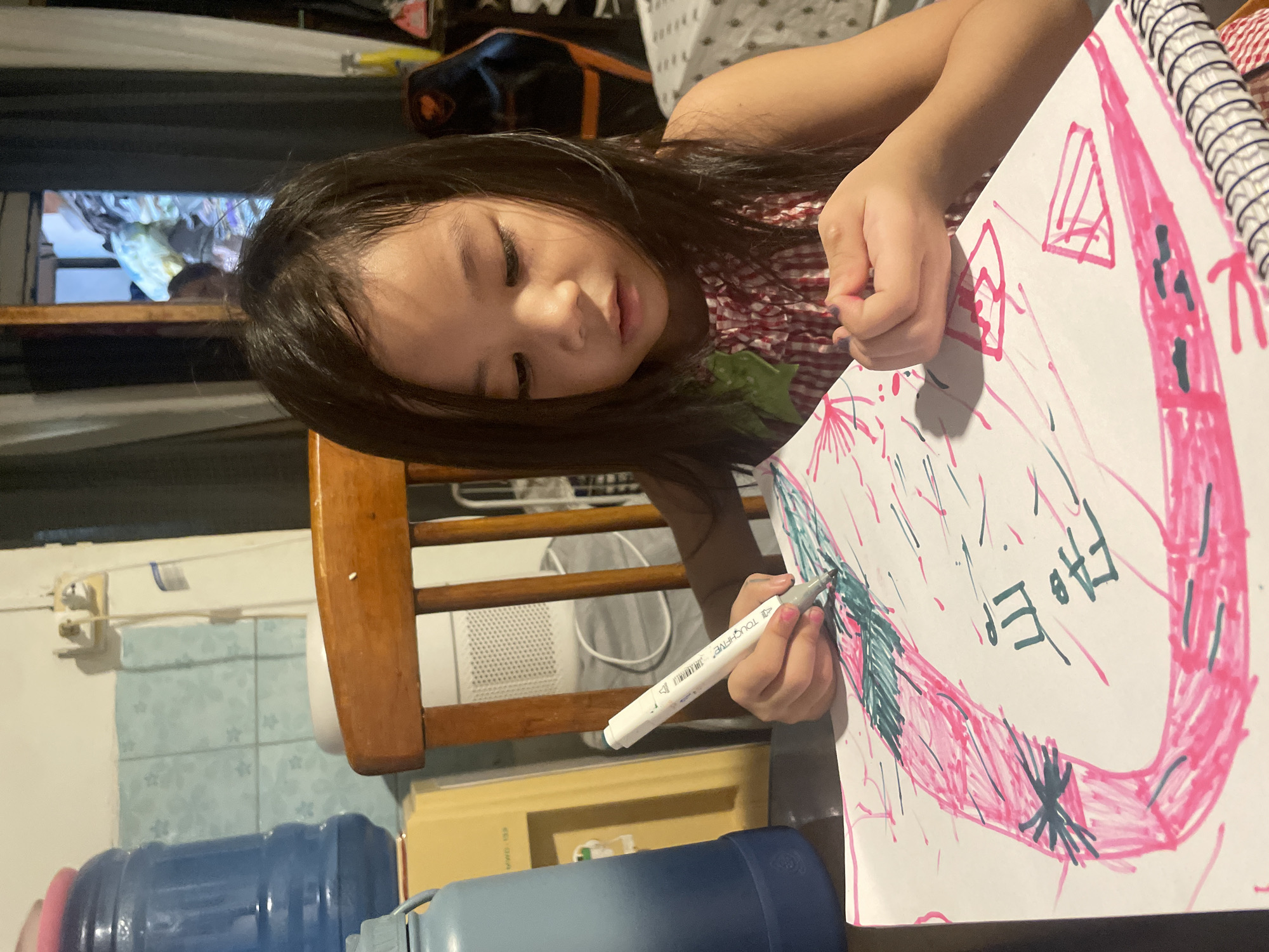 A young girl with dark hair is drawing on a large white paper pad. She is wearing a red and white checkered sleeveless dress and is sitting at a wooden table, holding a white marker in her right hand and coloring a drawing with pink and green markers. The drawing on the paper appears to be abstract, with bold pink lines and some green shading. There is text in what appears to be Korean written in black marker on the paper. The girl's left hand is resting on the paper, supporting it.
The environment appears to be indoors, possibly a home. Visible in the background are a large blue water dispenser, a wooden chair, a white air purifier, and a window with dark curtains. In the reflection of the window, a blurry image of another person can be seen. The lighting suggests it is daytime or evening.
The image's metadata indicates it was taken in Davao City, Philippines. Based on the visible elements like the water dispenser common in many Philippine households and the style of the interior, the scene is consistent with a residential setting in Davao City. The presence of common household items suggests a domestic environment rather than a public space.
