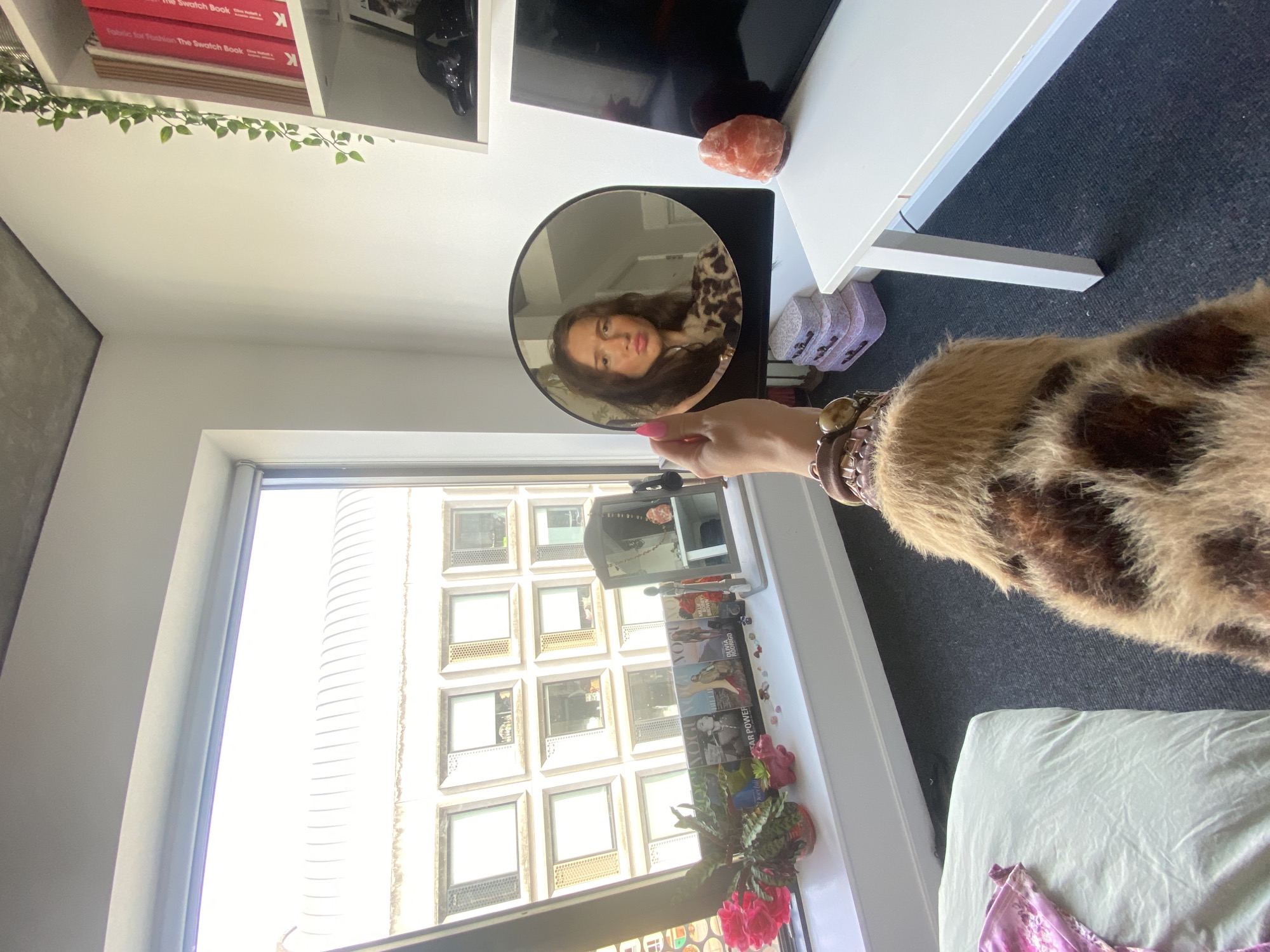 A young woman is holding a round mirror and looking into it. She has long brown hair and is wearing a fluffy, animal-print coat. She is making a pouty face and has a bright pink fingernail visible on her left hand. In the mirror, her reflection shows her face and shoulders. The mirror is placed on a white shelf unit.
The background shows a window with a view of modern buildings. There are books on shelves to the right of the mirror, and a pink Himalayan salt lamp on the shelf below. A black television screen is also visible. On the windowsill, there are magazines, a potted plant with pink flowers, and various decorative items. A bed with a light green duvet is in the foreground on the left. The room appears to be a bedroom. The overall impression is a personal, lived-in space.