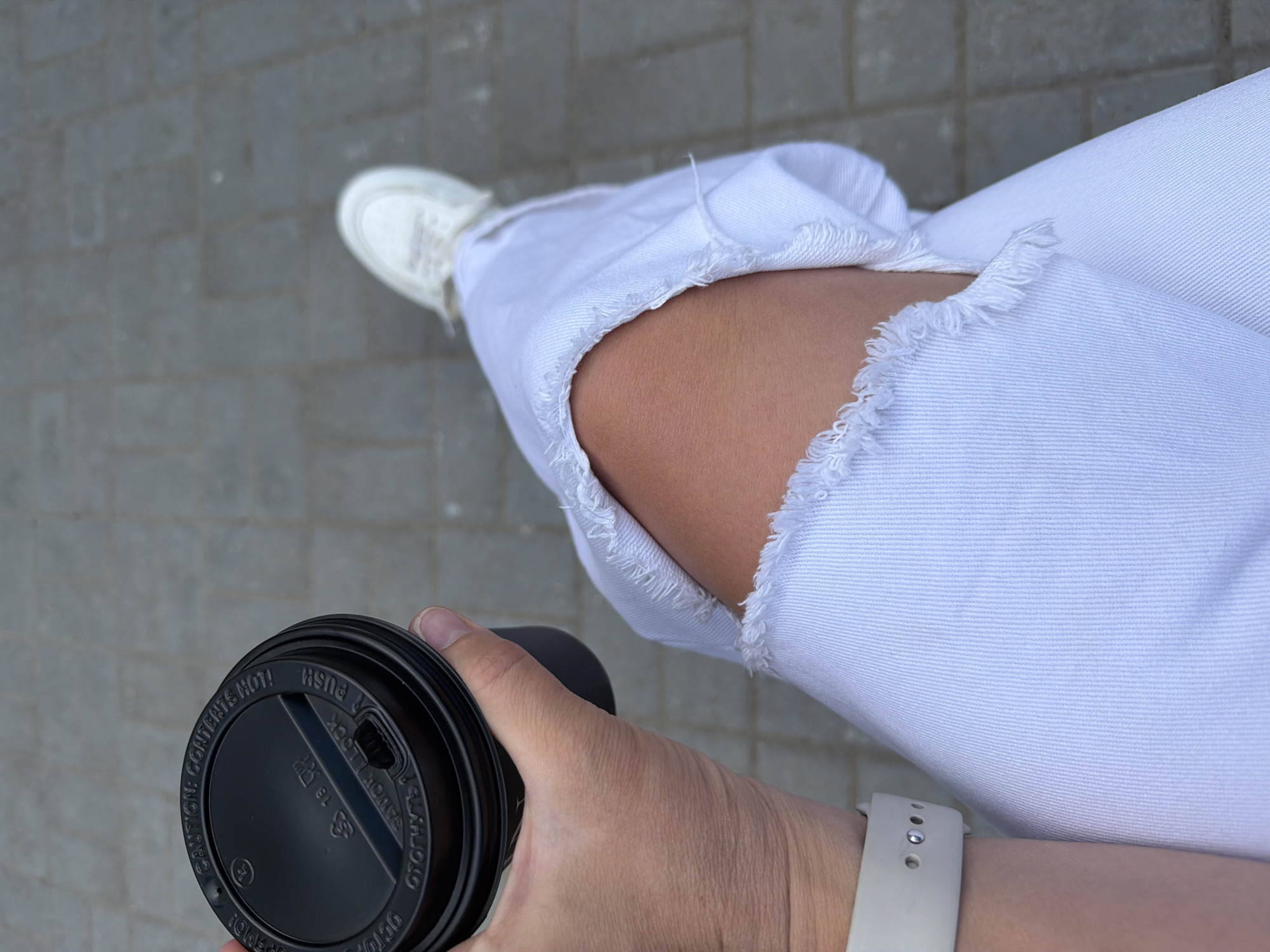 The image is a top-down shot showing a person holding a coffee cup while seated. The person wears white ripped jeans exposing their knee and white sneakers. A white smartwatch is visible on their left wrist. The coffee cup is black with a black lid that reads "CAUTION: CONTENTS HOT!" The ground is made of gray brick. The location is Chaikovsky, Russia.