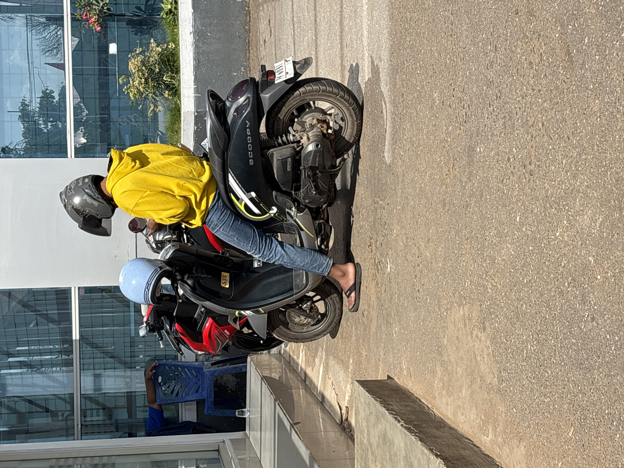 In Merauke, Indonesia, a person wearing a yellow hoodie and blue jeans sits on a black Honda Scoopy scooter. They are wearing a grey helmet with a Red Bull logo and a face shield. Another blue helmet is visible on the handlebars of the scooter. The scooter is parked on an asphalt road in front of a building with large glass windows that reflect trees and the sky. A person in a blue shirt is seated in a blue plastic chair to the left of the frame, near a glass of water. The environment appears to be a sunny day, with shadows cast on the ground.
