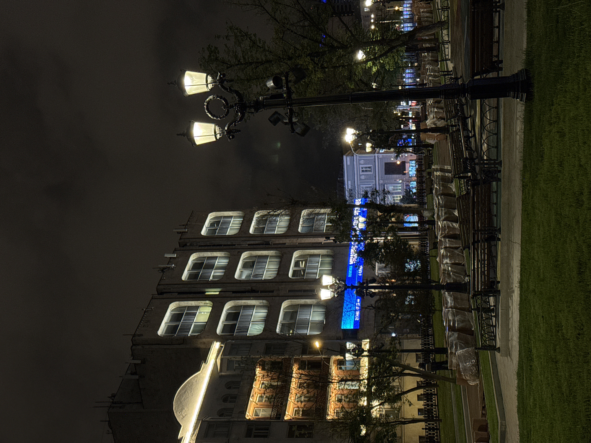 The image depicts a nighttime scene in Moscow, Russia. A multi-story building with lighted windows is on the left. Streetlights illuminate the scene. On the right is a park-like area with covered benches. The sky is dark and overcast.