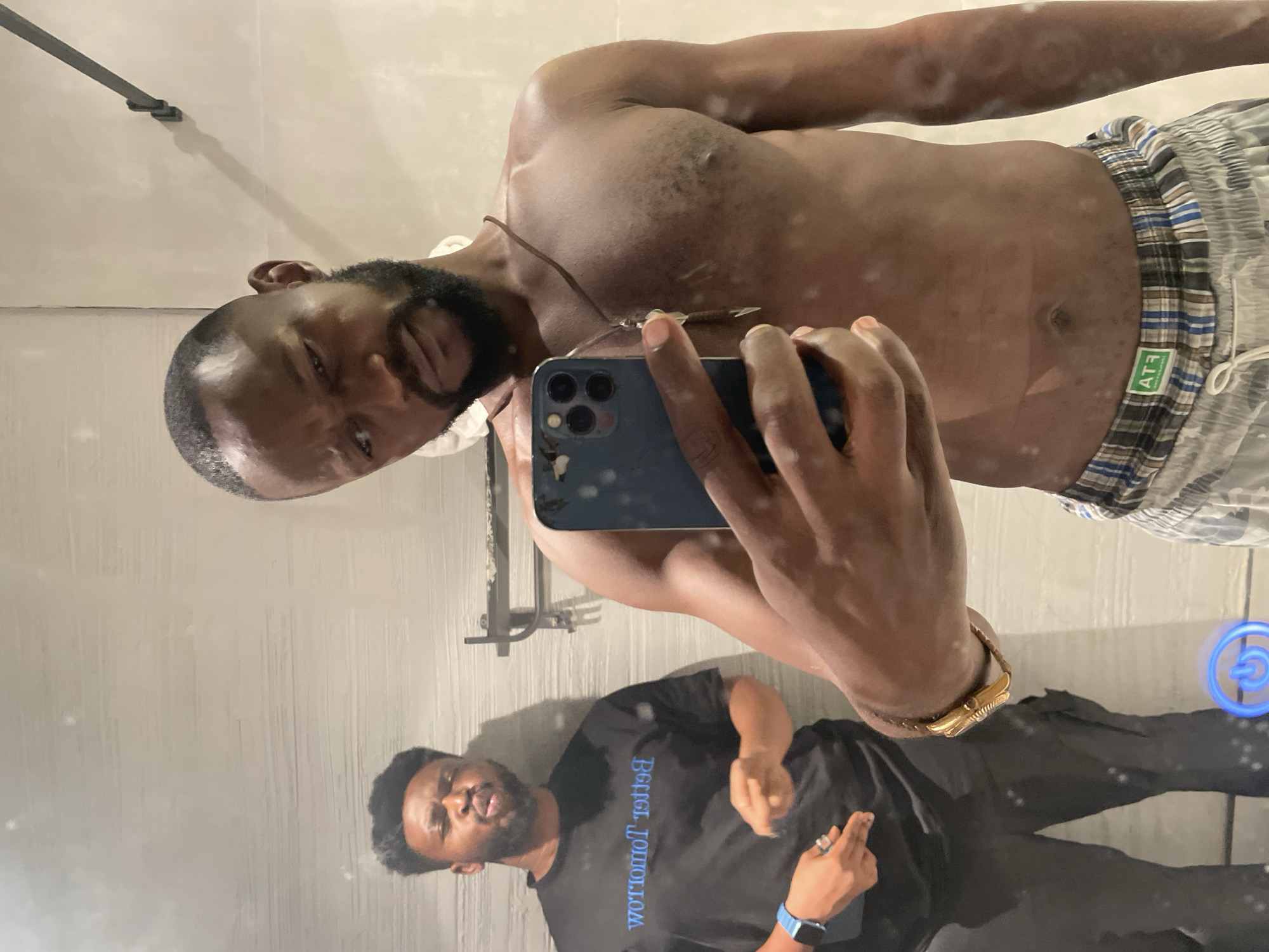 The image is a mirror selfie taken indoors, rotated 90 degrees counter-clockwise from its natural upright orientation. It features two dark-skinned men, likely in a bathroom or changing area with tiled walls, in Abuja, Nigeria.
The primary subject, centered and closer to the camera, is a shirtless man with a neatly trimmed beard and short hair. He is wearing blue, white, and brown plaid boxer-style shorts with a small "AT3" tag visible on the waistband. He holds a blue smartphone, likely an iPhone, in his right hand, capturing his reflection. A brown beaded necklace with a metallic pendant hangs around his neck, and a gold-toned watch adorns his left wrist. His expression is neutral as he looks directly into the mirror.
In the background, further back in the reflection, stands a second man. He also has a beard and is wearing a black t-shirt with the reversed text "worromot retteB" (Better Tomorrow) prominently displayed. He is also wearing black pants and has a blue smartwatch on his left wrist and a ring on his right hand. He is making a gesture with his right hand, possibly an inverted middle finger.
The mirror surface shows noticeable smudges and marks. A black metal bar, possibly a towel rack, is visible on the wall in the reflection. A faint blue glowing circular object with a power symbol is also visible in the bottom right of the reflection. The lighting is indoor and appears to be artificial.