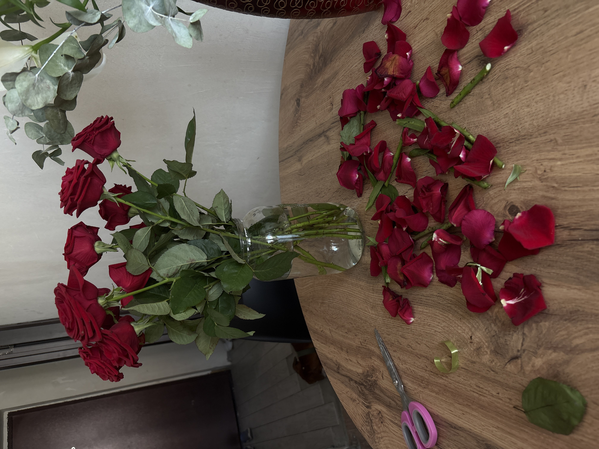 A bouquet of red roses in a glass vase sits on a wooden table. Scattered around the vase are rose petals and leaves, along with a pair of pink-handled scissors and a piece of green ribbon. In the background, a door is visible. The scene appears to be an indoor setting, possibly a home in Rostov-on-Don, Russia.