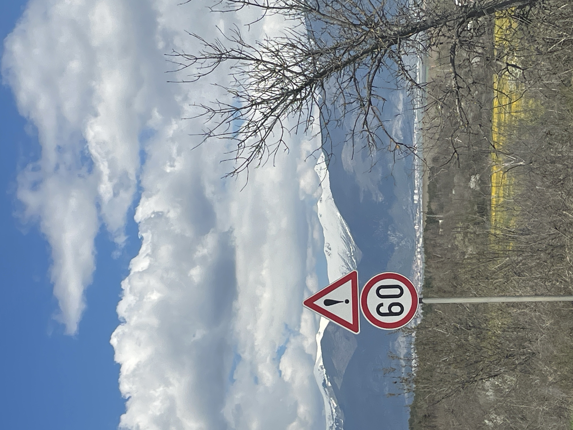 The image shows a landscape scene. In the foreground, a road sign with a triangular warning symbol and a circular sign indicating a speed limit of 60 are visible. Behind the sign are snow-capped mountains that dominate the background. In the sky, there are fluffy white clouds, contrasting with patches of blue sky. The terrain consists of vegetation and trees, partially obscuring a stretch of yellow field. There are no people visible in the scene. It's impossible to verify the location as Mihiltsi, Bulgaria.