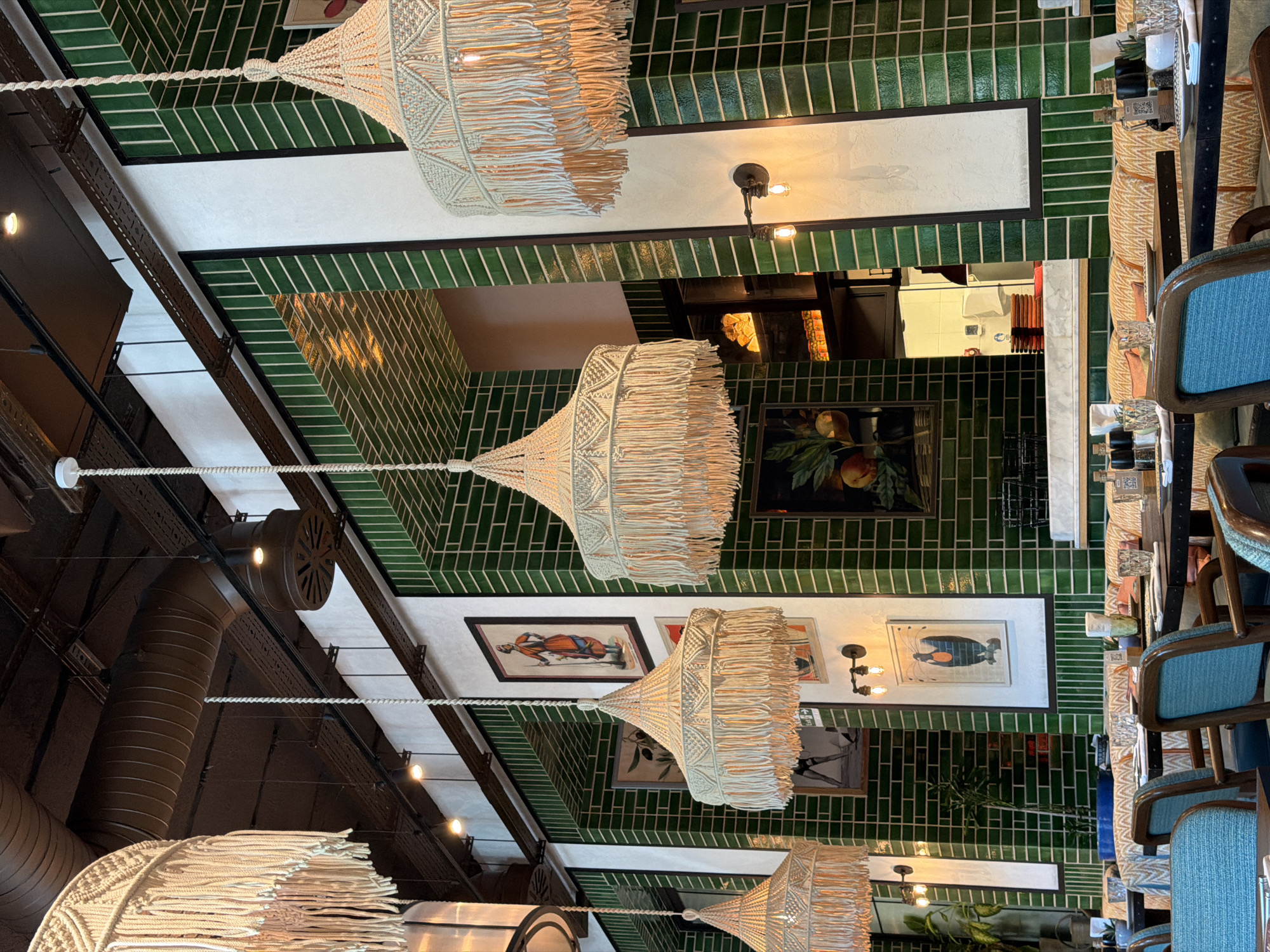 This is an interior shot of a restaurant in Sabail Raion, Azerbaijan. The space features high ceilings with exposed industrial ductwork and dark wooden beams. Several large, macrame pendant lamps with fringe hang from the ceiling, casting a warm glow. The walls are predominantly covered in glossy, dark green tiles arranged in a vertical brick pattern, punctuated by white rectangular panels. Framed artwork depicting figures and still life hangs on the tiled walls.
The foreground shows a dining area with dark tables set with white placemats, glassware, and salt and pepper shakers. The chairs have blue upholstered seats and backs with dark wooden frames. Behind the tables, banquettes with patterned upholstery line the walls. In the background, a glimpse into the kitchen area reveals shelves stocked with food items and white tiled walls. The overall ambiance suggests a trendy and stylish dining establishment. There are no people visible in the frame.
