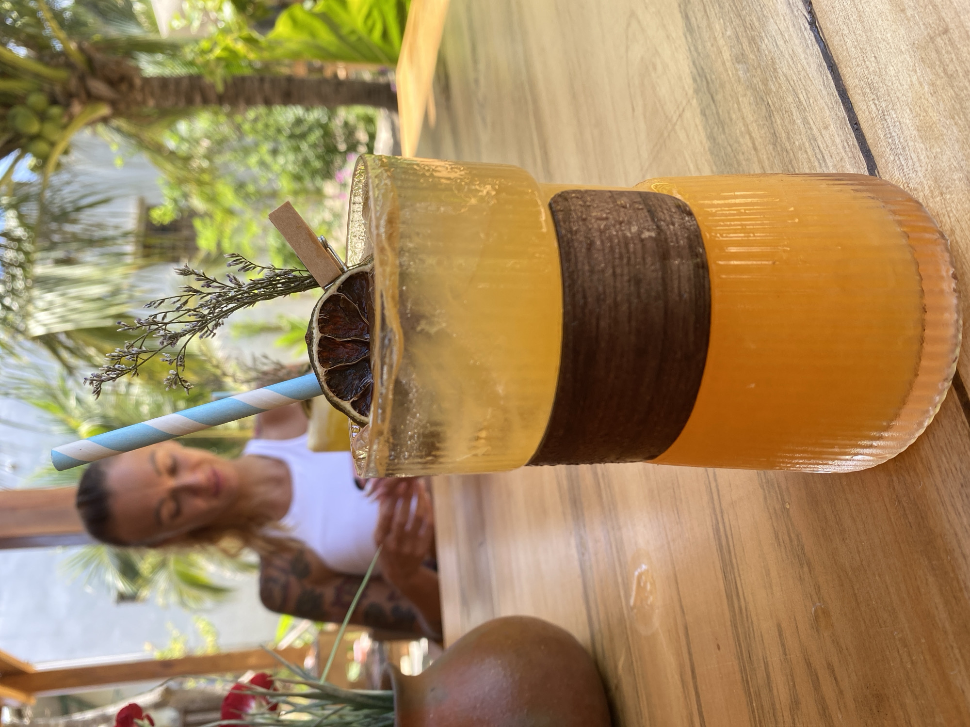 In Phu Thuy Ward, Vietnam, a close-up shot focuses on a tall, clear glass filled with an amber-colored beverage, possibly a cocktail or juice, adorned with a slice of dried lime, a sprig of dried foliage, and a small wooden clip holding the foliage in place. A blue and white striped straw protrudes from the drink. The glass is multifaceted and has a dark brown woven band around its middle. It sits on a wooden table, which shows water rings and streaks. In the background, out of focus, a woman wearing a white tank top with tattoos on her left arm is seated. To the left of the glass, a small, brown, earthenware vase holds red flowers. The setting appears to be outdoors, with palm trees and lush greenery visible in the background under a bright sky.