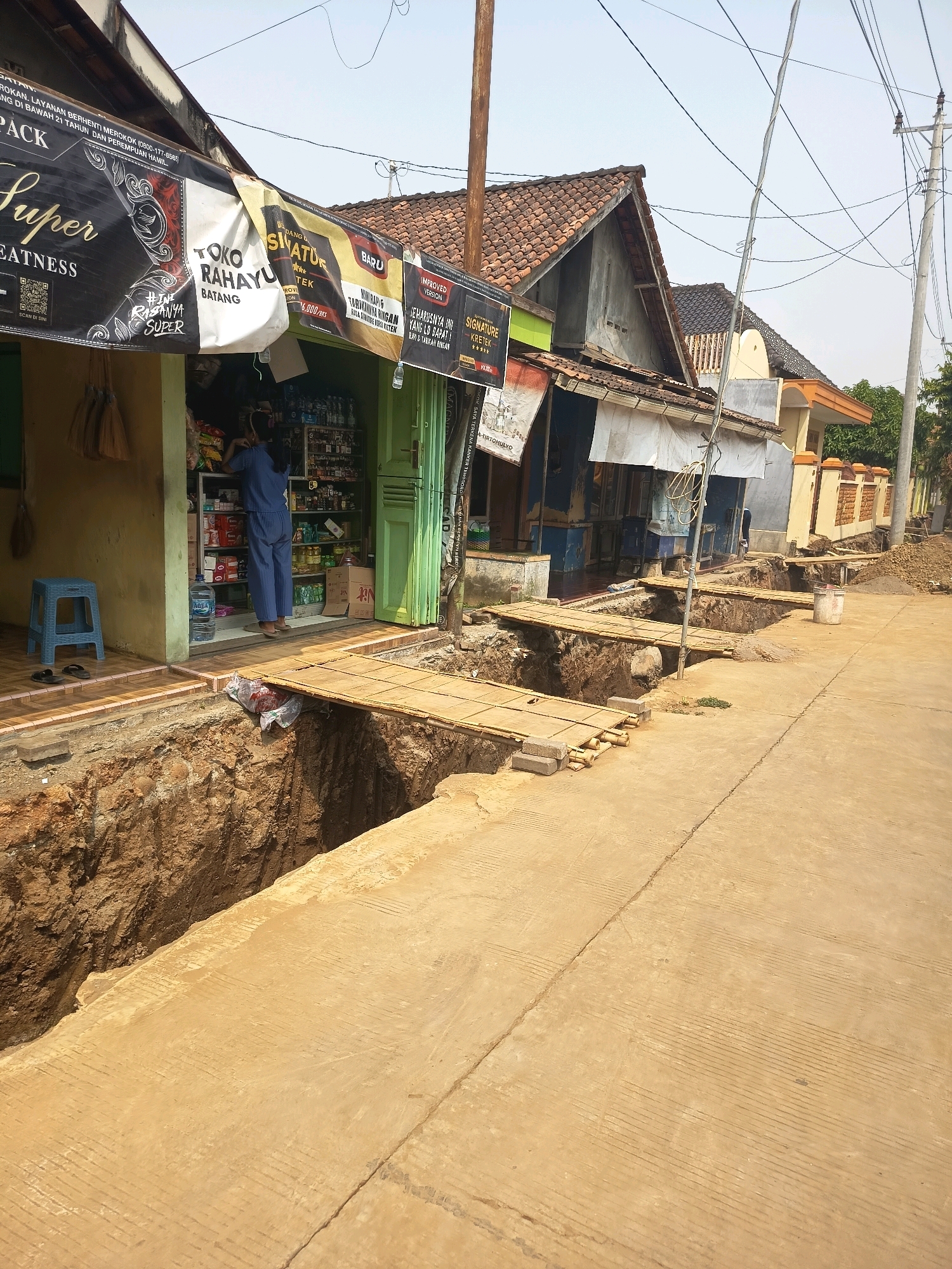 The image depicts a street in a state of disrepair, with a large section of the road collapsed, creating a deep trench. Temporary bridges made of wood planks have been constructed across the trench to allow passage. The scene shows a row of small shops or houses lining the street, some with awnings and signage. In the foreground, a person in blue clothing is standing inside one of the shops, appearing to be attending to shelves of goods. A blue plastic stool and a pair of sandals are visible outside the shop. The environment suggests a dry, sunny day. The overall impression is one of a community adapting to a significant infrastructure issue.