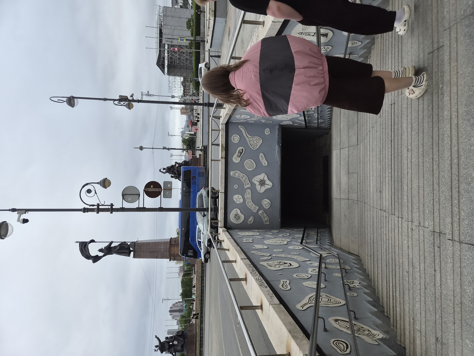 This is a street-level photograph taken in Vladivostok, Russia, featuring a pedestrian underpass and its immediate surroundings. In the foreground, a wide, gray concrete staircase leads down into the underpass. The walls of the underpass are decorated with a stylized mural featuring abstract shapes and figures reminiscent of fossils or sea creatures, rendered in white outlines on a gray background. Metal railings line the sides of the staircase.
Two individuals are partially visible on the staircase. The person in the foreground, viewed from behind, is wearing a pink and black jacket, dark brown shorts, and white socks with striped sandals. To their right, another person is visible from the chest down, wearing dark pants and white socks with white slip-on shoes.
Beyond the underpass and staircase, a street is visible with a blue bus and two cars. In the background, several large statues are prominently displayed, including a prominent one of a soldier holding a flag. Streetlights and traffic signals are also present. The sky is overcast and gray, suggesting a cloudy day. The overall scene depicts a functional urban space with artistic embellishments, a common feature in public pedestrian areas.