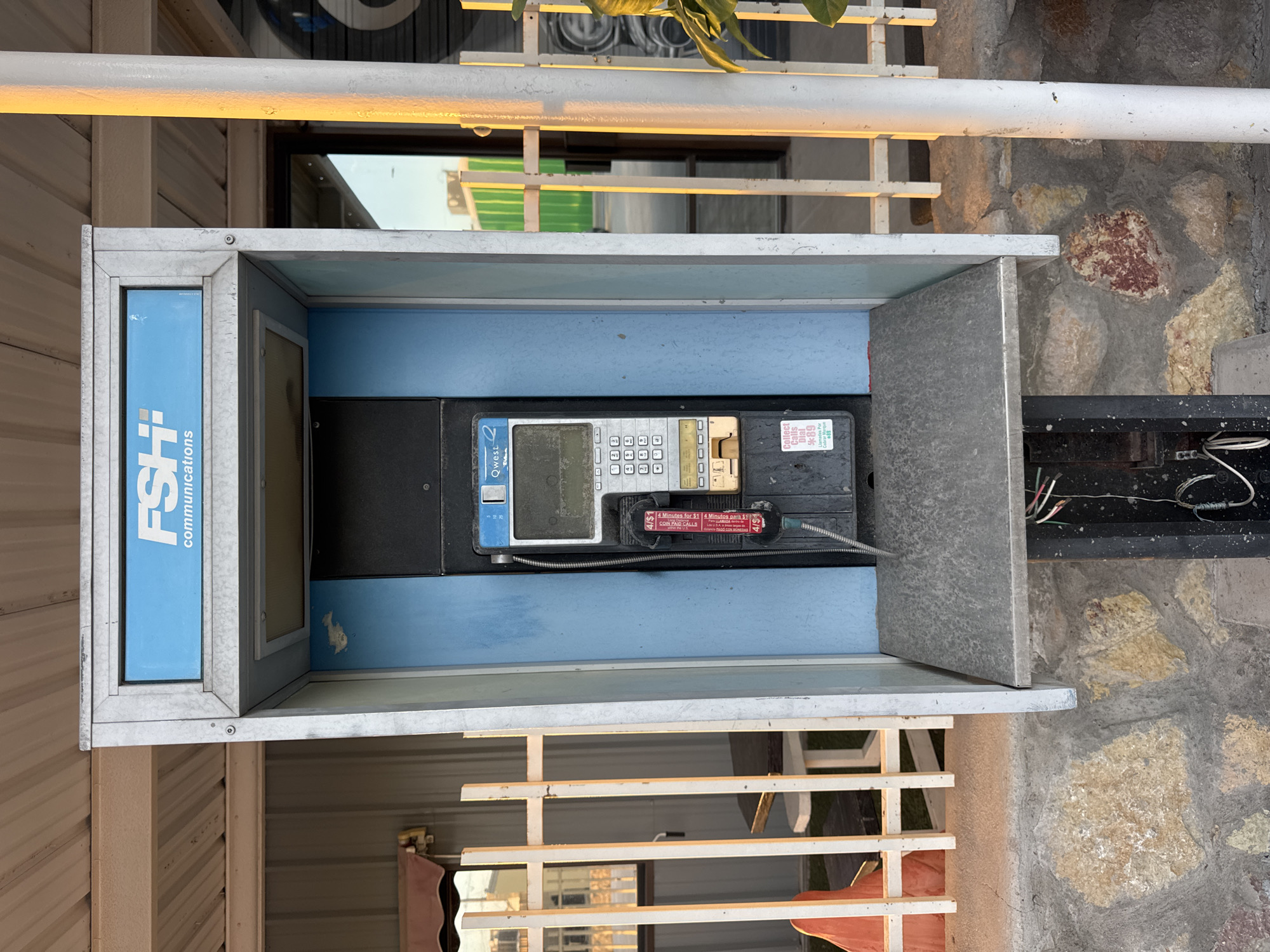 A public payphone booth, likely located in Sunland Park, New Mexico, United States of America, stands in front of a building with a corrugated metal exterior. The booth itself is constructed with a galvanized metal frame and blue panels. At the top of the booth, a sign displays "FSH Communications" in blue lettering on a white background. Inside the booth, a payphone unit is mounted. The phone features a dark gray casing with a digital display screen and a keypad. A black handset is attached by a coiled cord. Below the keypad, there is a coin slot and a card reader. A sticker on the lower part of the phone reads "Collect Calls Dial *89". The payphone booth sits on a stone foundation, and the area around it appears to be outdoors. A white metal fence runs along the side of the booth, and in the background, a building with green siding and reflections of the sky can be seen. The lighting suggests it may be late afternoon, with warm sunlight illuminating the scene.