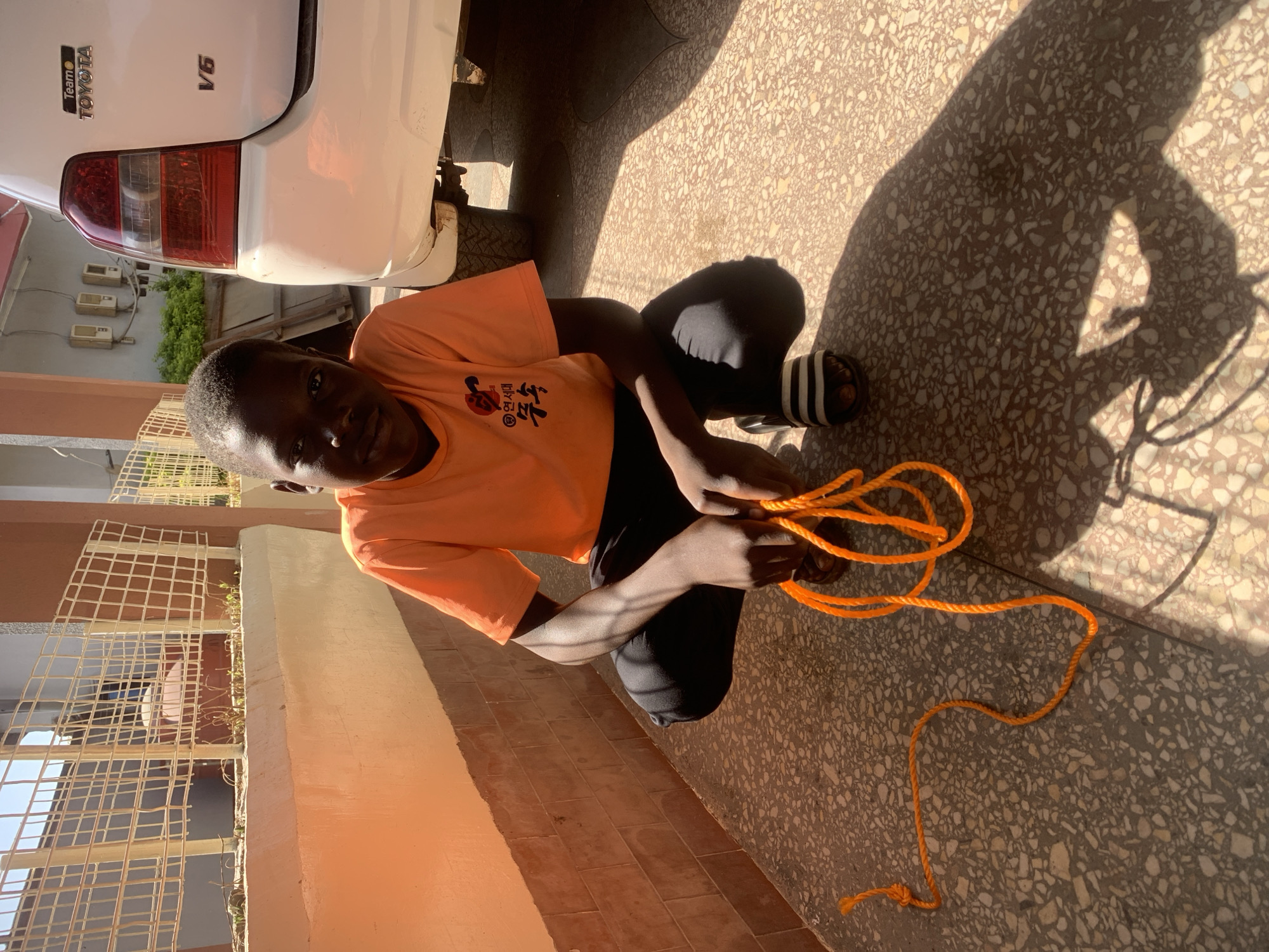 A person is sitting on the ground holding an orange rope. The person is wearing an orange shirt with black pants and black and white sandals. A white Toyota vehicle is visible in the upper left corner of the image. The environment consists of a tile ground and a building with a caged window. Based on the context provided, the location is likely in the city of Jos, Nigeria.