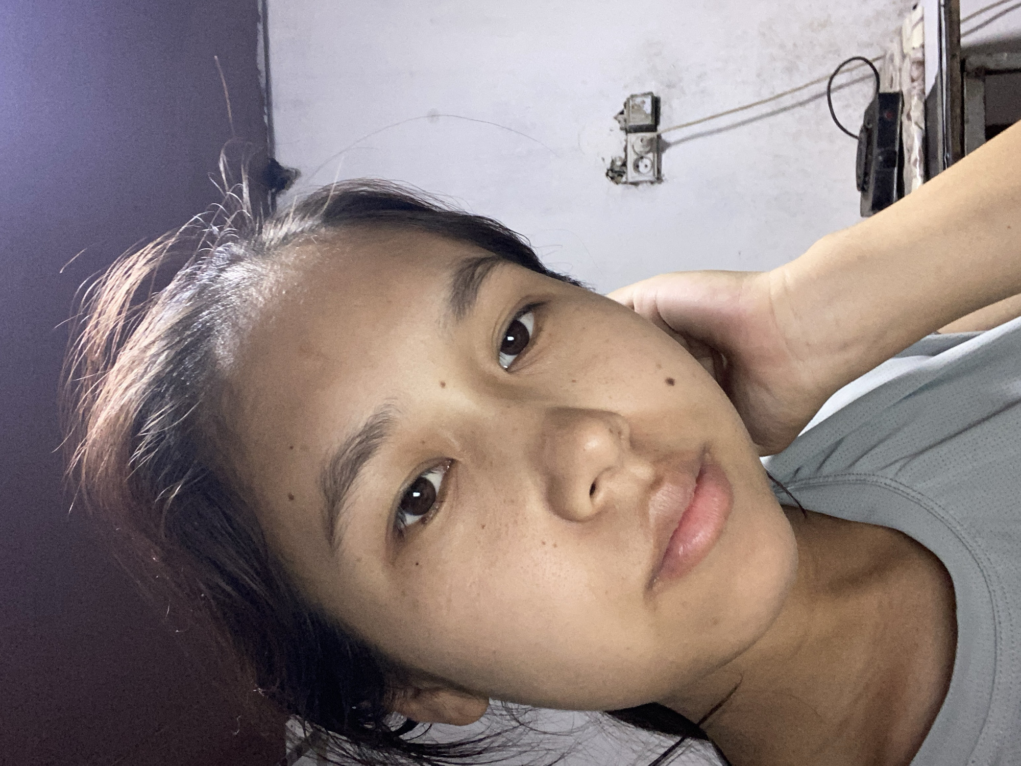 The image is a close-up, slightly overhead selfie of a young woman. She has dark hair, pulled back loosely, with some strands framing her face. Her skin is light brown with visible freckles and a few moles. She is wearing a light grey t-shirt. Her left hand is resting on her cheek and jawline, and her right arm is bent and supporting her head. Her expression is neutral, with her eyes looking directly at the camera. In the background, a white wall is visible, with a white electrical outlet and some wires mounted on it. A black electronic device with a coiled cord sits on a table or surface in the lower right corner. The lighting suggests an indoor setting, possibly at night or in a dimly lit room. There is no indication of the specific location within Dostuk, Kyrgyzstan.