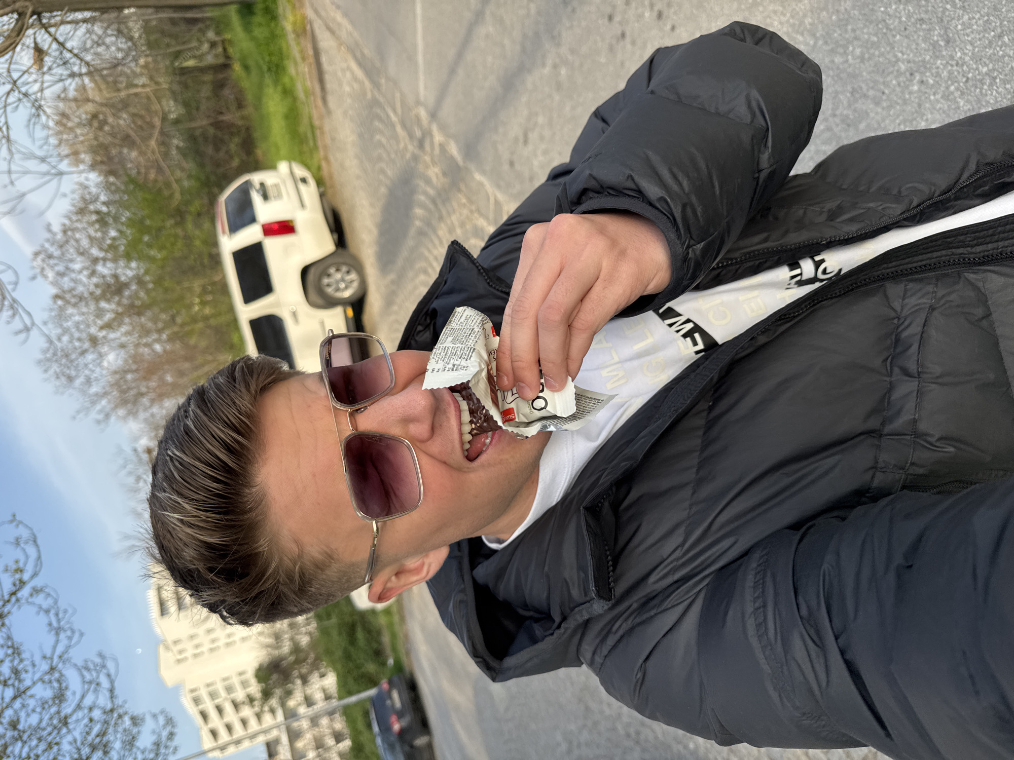 A man is seen in an outdoor selfie taken in Plovdiv, Bulgaria, eating a chocolate bar. He has short, light brown hair and is wearing sunglasses, a black puffer jacket, and a white t-shirt. He holds the opened chocolate bar wrapper with his right hand. In the background, there's a white SUV with black details parked on a paved road. Trees, a building, and blue sky are visible in the distance. The photo appears to be taken on a city street, likely during daylight.