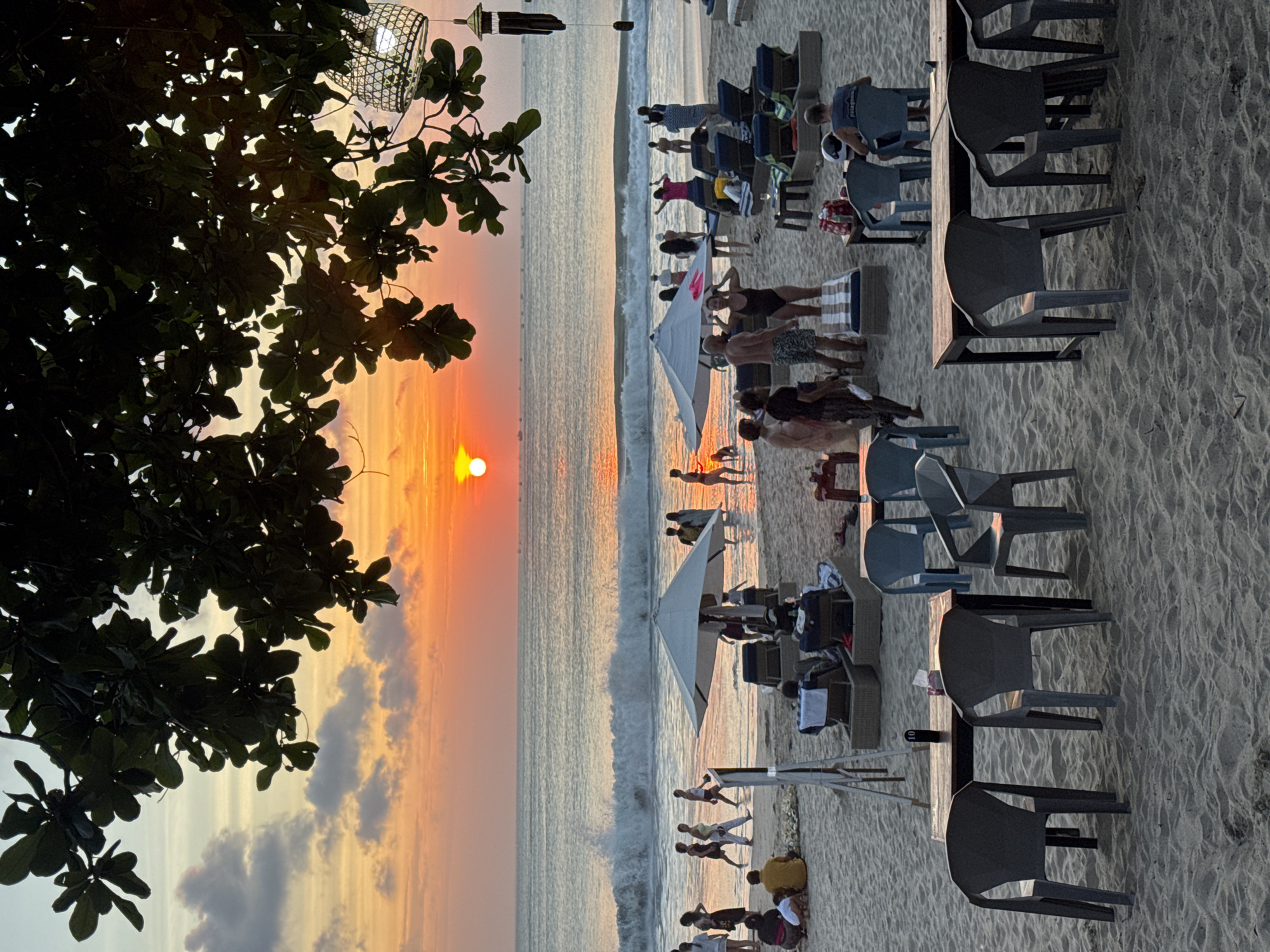The image depicts a serene sunset scene on a beach in Kuta Selatan, Indonesia. The sky is painted with vibrant hues of orange and yellow, with the sun partially visible as it descends towards the horizon. Gentle waves roll onto the sandy shore, where several people are enjoying the evening. Some individuals are strolling along the water's edge, while others are relaxing on beach chairs or under umbrellas. In the foreground, tables and chairs are set up on the sand, suggesting a dining area or lounge. The overall atmosphere is tranquil and picturesque, capturing the essence of a tropical beach at sunset.