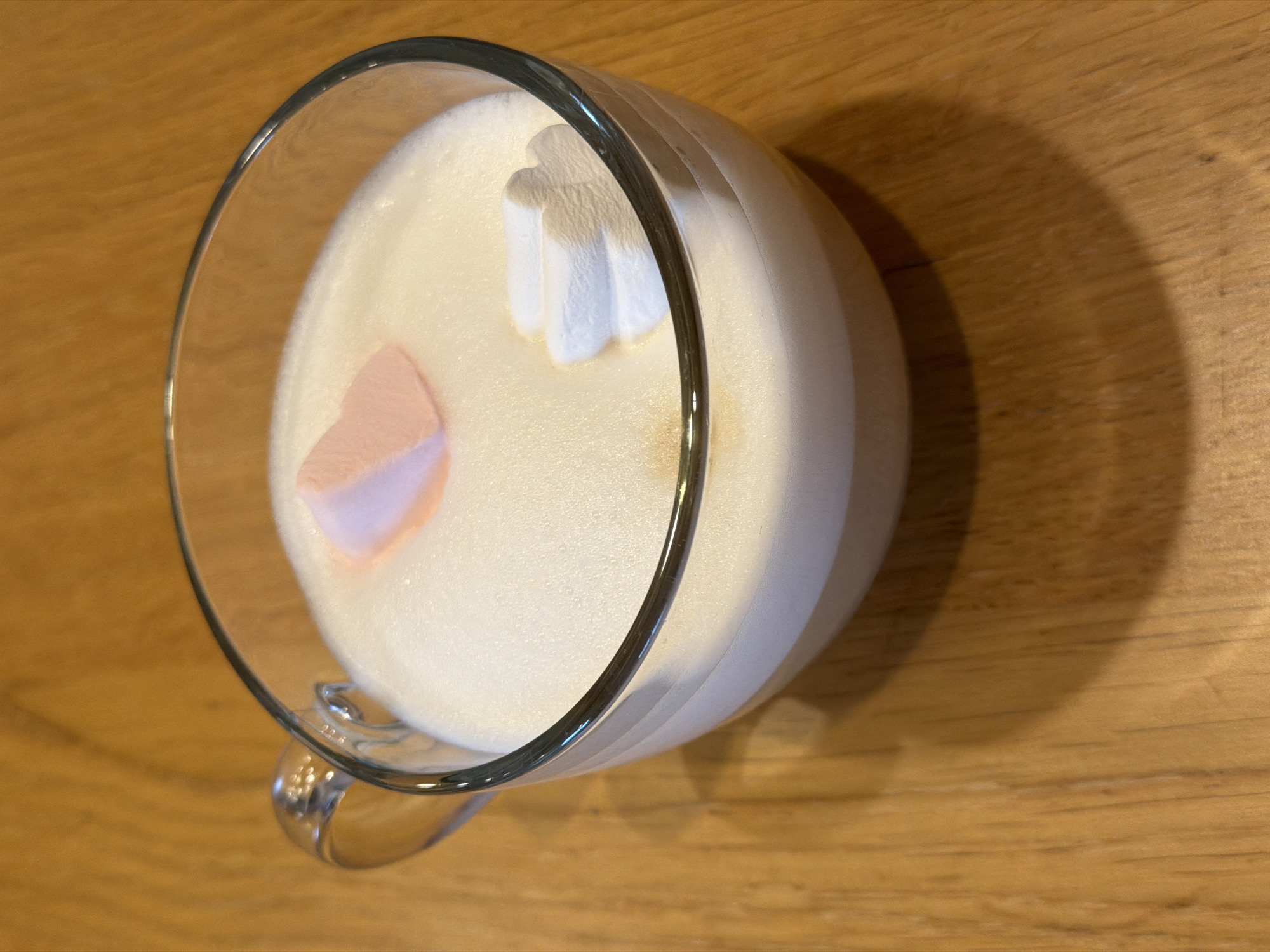 A glass mug of frothy, light brown beverage is placed on a wooden table. The mug has a handle and is filled to the brim with the drink, topped with a thick layer of foam. Two marshmallows, one pink and heart-shaped, and another white and rectangular, are floating on the foam. The table has a visible wood grain texture and a warm, brown tone. The lighting suggests an indoor setting, possibly a kitchen or cafe, but no specific city or location details are discernible.