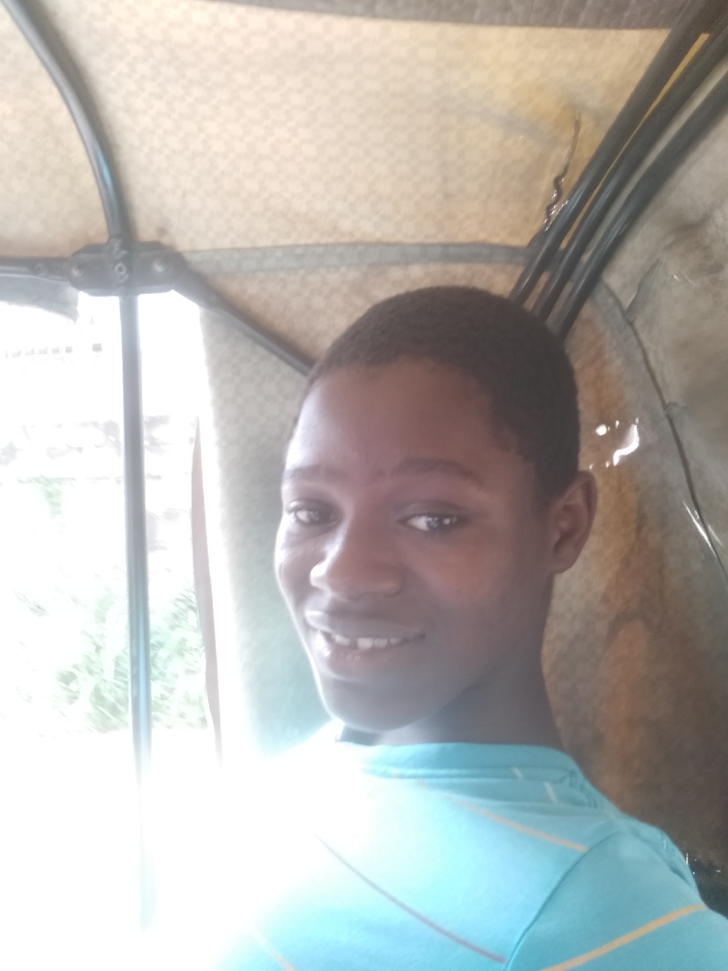 A close-up, eye-level shot shows a young Black male inside an auto-rickshaw (tuk-tuk). He is smiling, wearing a light blue t-shirt with thin yellow and brown stripes. The vehicle's interior is visible, including parts of the metal frame, the roof lining with a patterned design, and clear plastic sheeting on the sides. Outside the open window on the left, blurred greenery and a building are partially visible. It is presumed that this is in Shangisha, Nigeria.