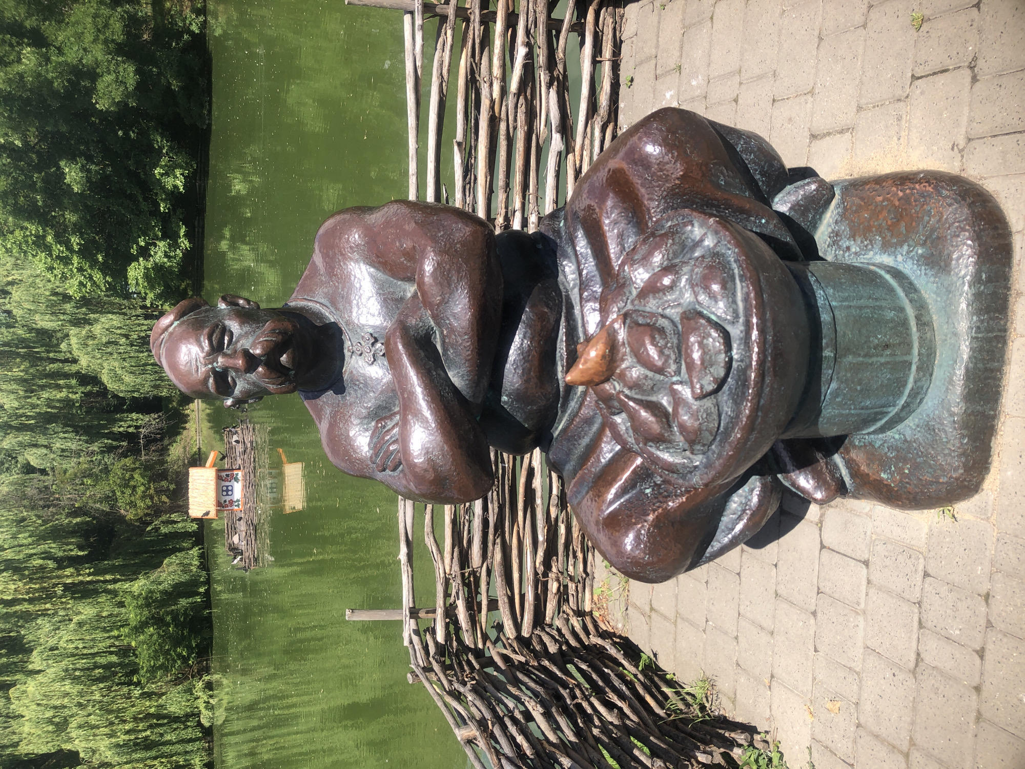 This is a medium shot, eye-level, outdoor photograph of a bronze statue of a Cossack in a seated position. The statue is positioned in the foreground, facing forward, with its arms crossed and its head tilted slightly downwards. The statue is dark brown with green patina in some areas. The Cossack has a prominent mustache and is wearing a small cap on his head. He is seated on a cylindrical base, and in front of him, resting on his lap, is a bowl filled with what appear to be fruits or vegetables.
Behind the statue, a body of water, likely a pond or lake, is visible. The water is a vibrant green color, and it reflects the lush, green foliage of trees that surround the body of water. The trees are dense and form a natural backdrop to the scene. In the middle of the water, a small, wooden structure resembling a miniature house or gazebo is floating. This structure has a thatched roof and is supported by a raft-like base.
A rustic fence made of interwoven branches separates the statue's immediate area from the water. The fence is low and appears to be made of natural, unfinished wood. The ground in front of the statue is paved with light-colored cobblestones. The overall lighting suggests a sunny day, with bright sunlight casting shadows. The scene evokes a tranquil park or garden setting.