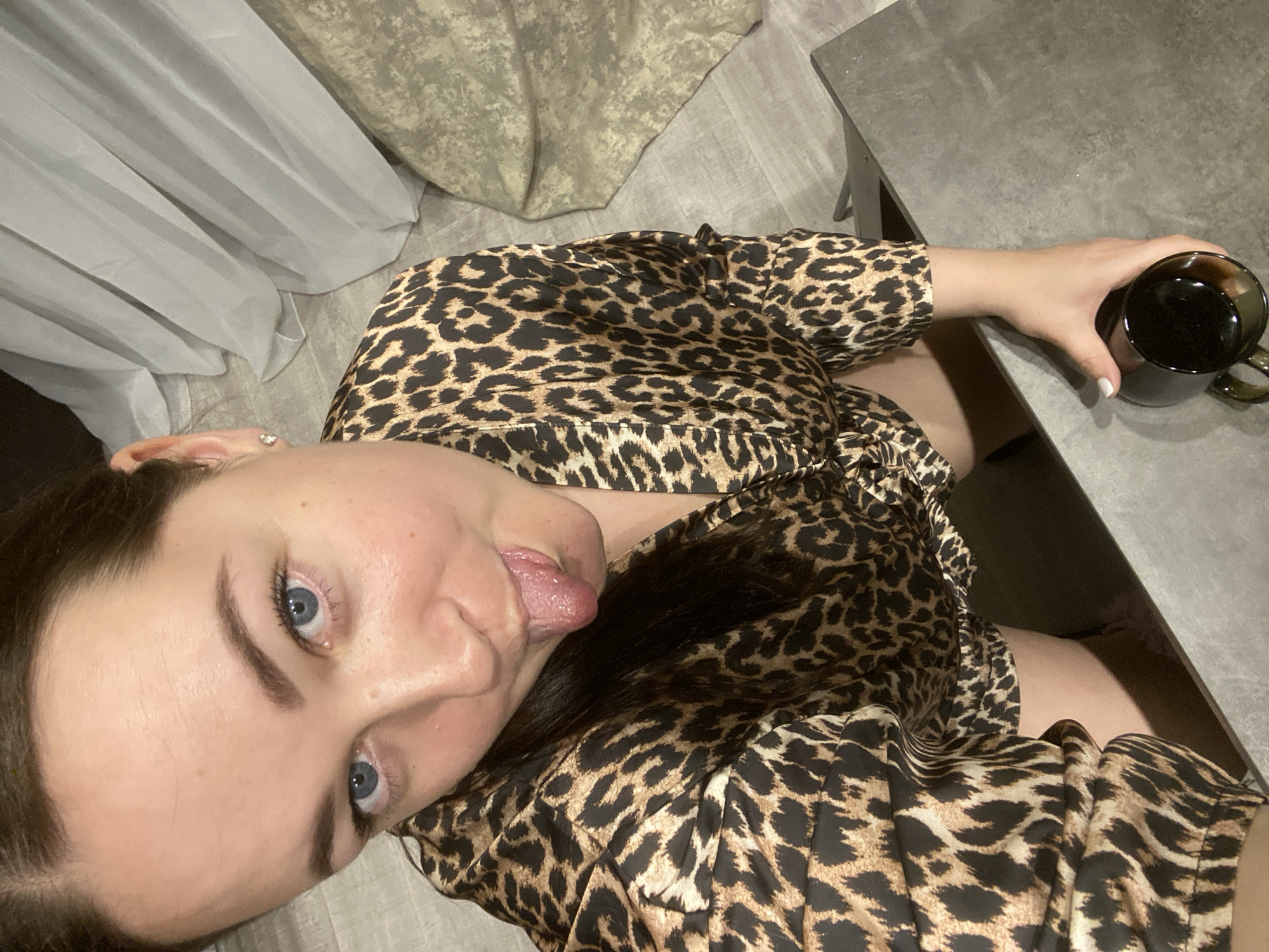 A woman with blue eyes and dark hair is lying on her side, wearing a leopard print robe. She is sticking her tongue out and looking at the camera. She is holding a dark-colored mug on a gray table. In the background are white and tan curtains. The location is not discernable from the provided image.