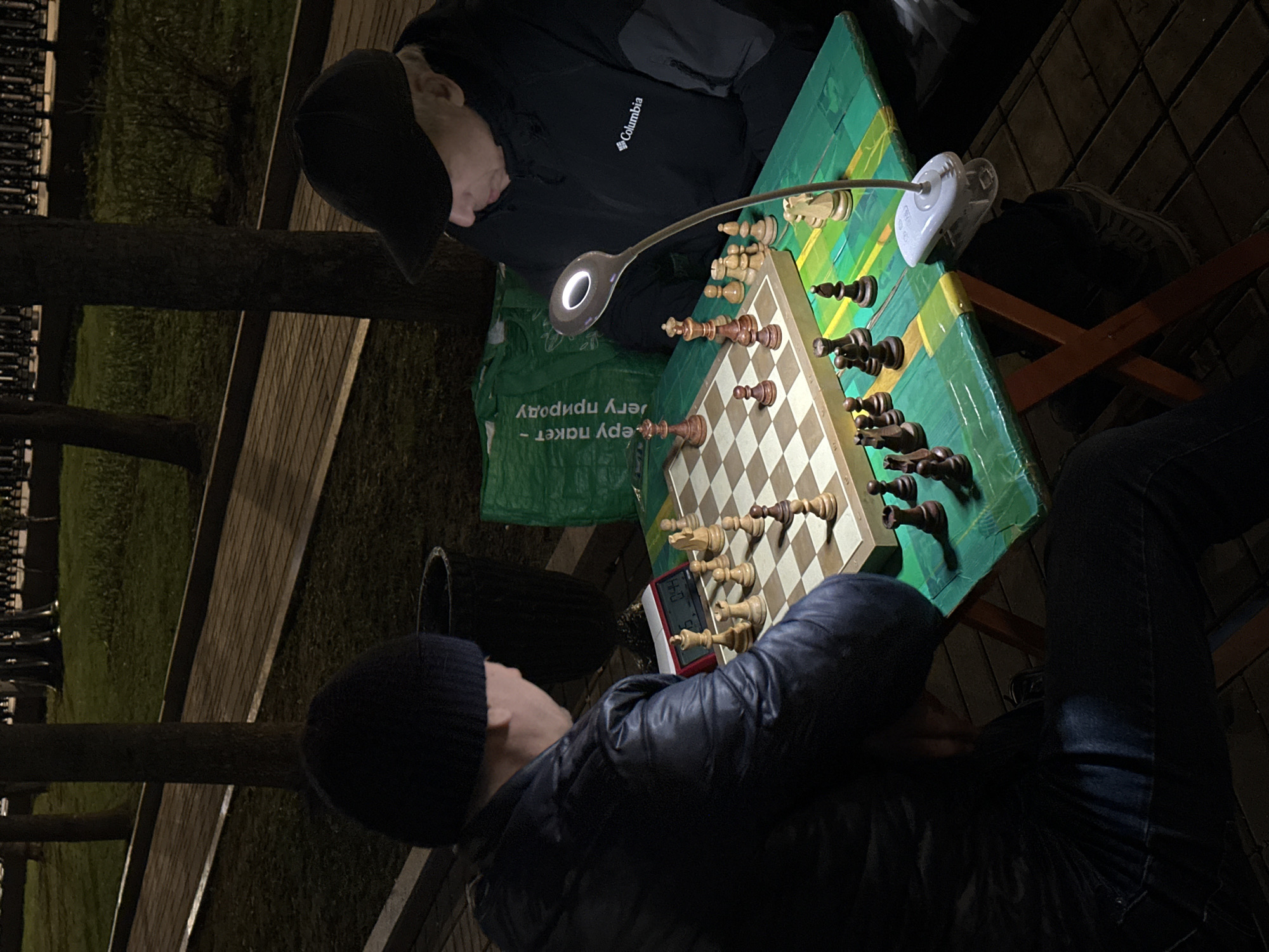 Two people are engaged in a game of chess outdoors at night. The chess board is lit by an adjustable lamp. One person is wearing a black Columbia jacket and a baseball cap. The other person is wearing a black beanie and a dark puffer jacket. A chess clock is visible on the table. A green reusable grocery bag sits next to the chess board. The ground appears to be brick. In the background are trees, grass, a sidewalk, and a fence. The location is Moscow, Russia.