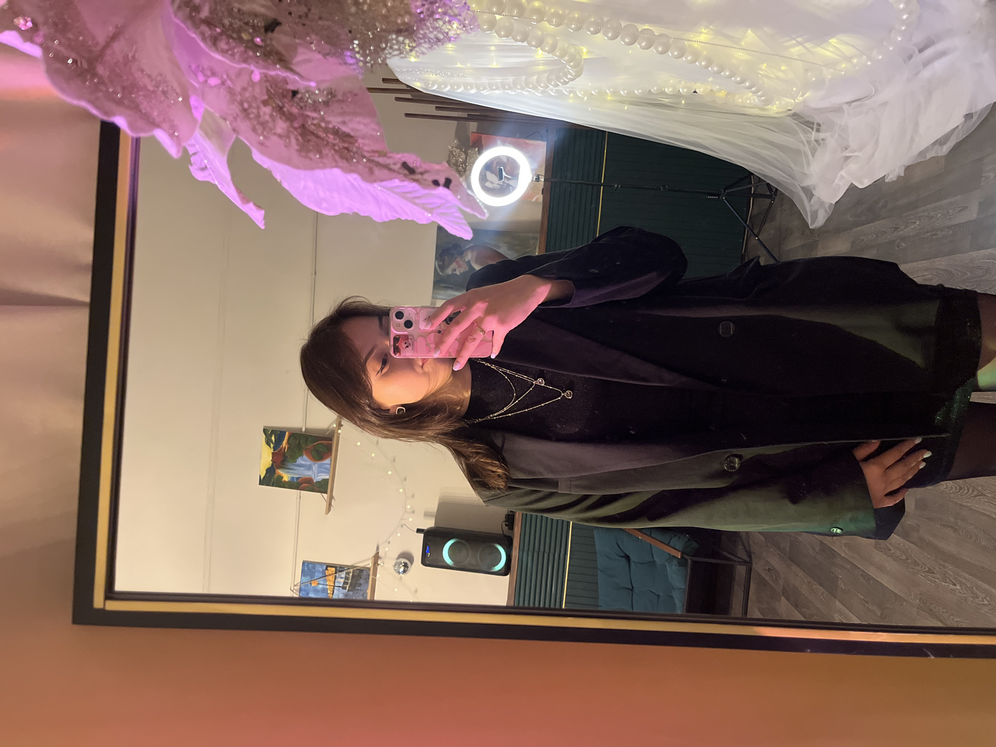 A young woman takes a mirror selfie with a pink cell phone. The reflection in the mirror shows her wearing a dark shirt and a green jacket. She also wears a necklace. Behind her is a wall with small paintings, a speaker, and a blue couch. Above her is a white, semi-transparent fabric with light-up string of pearls on it and a large pink flower. A circular light is also visible in the background. The location is likely indoors, but there is no information in the image about the city of Saint Petersburg, Russia.