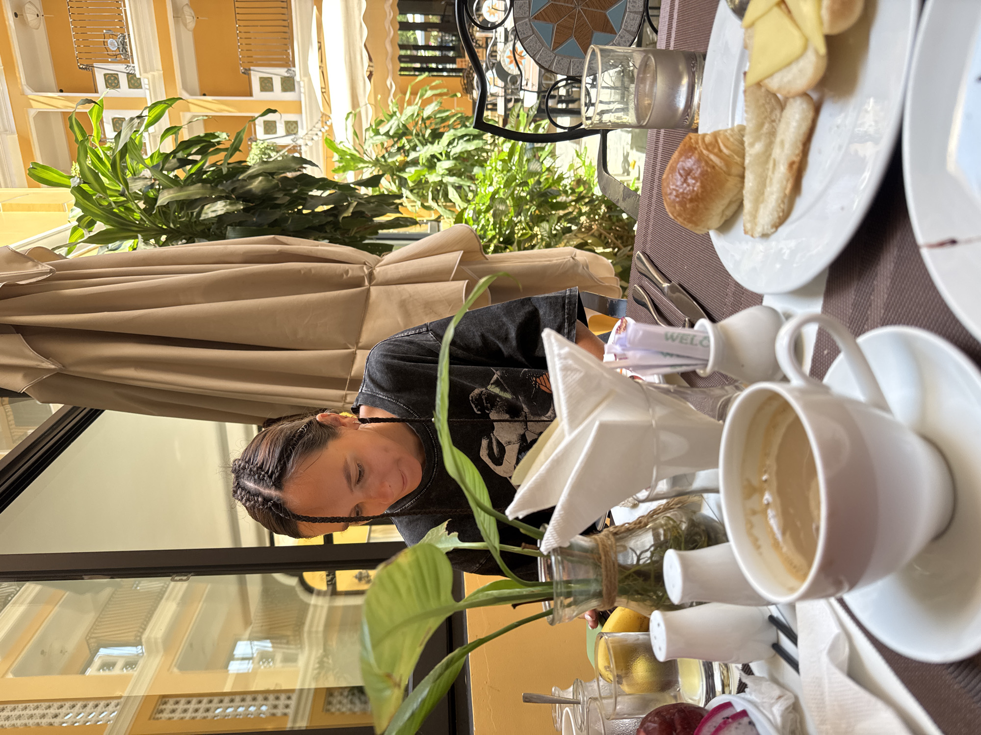 A woman with braided hair is sitting at a table in an outdoor dining area. She is wearing a black t-shirt. In front of her is a table with a brown tablecloth, a cup of coffee, a plate with bread and cheese, a glass of water, a small potted plant, sugar and creamer containers, and a napkin holder. The dining area is located outside a yellow building, possibly a hotel, in the city of Đà Nẵng, Vietnam. There is foliage behind the woman and the table is partially shaded by an umbrella.