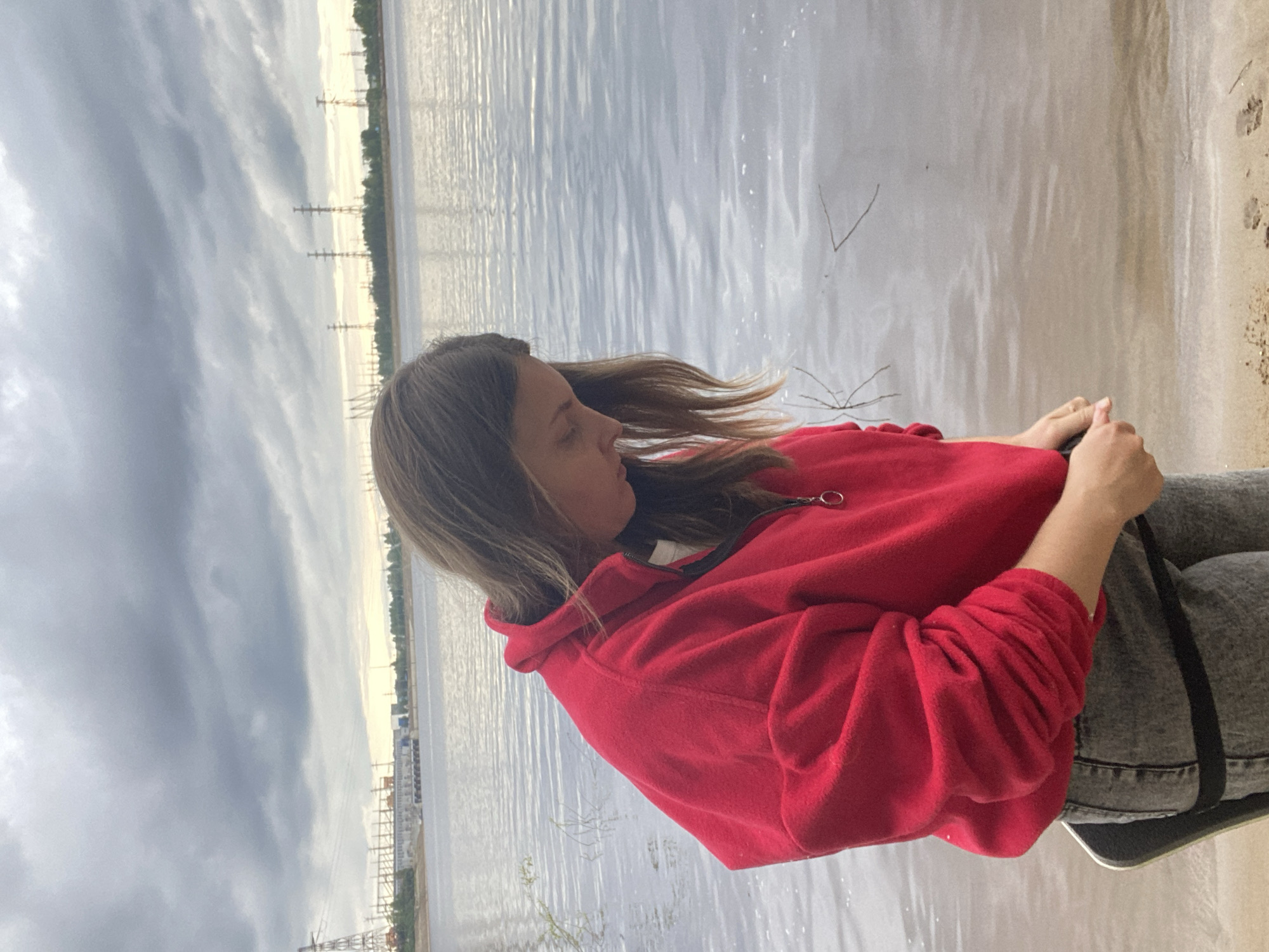 A young individual, likely female, with medium-length brown hair, stands by the sandy bank of a large body of water. She wears a red fleece half-zip pullover and grey denim jeans. Her head is bowed, and she looks down towards the water's edge, with her hands clasped or adjusting something at waist level. A black object, possibly a skateboard, is partially visible and strapped to her side. The water shows ripples and reflects the heavily overcast, grey sky. On the distant bank, a series of industrial structures are visible, including a large power transmission tower on the left, a complex of buildings resembling a hydroelectric power station, and a line of several power pylons extending across the horizon. This scene is consistent with the landscape along the Kama River in Chaikovsky, Russia, particularly with the Votkinsk Hydroelectric Power Station visible in the background.