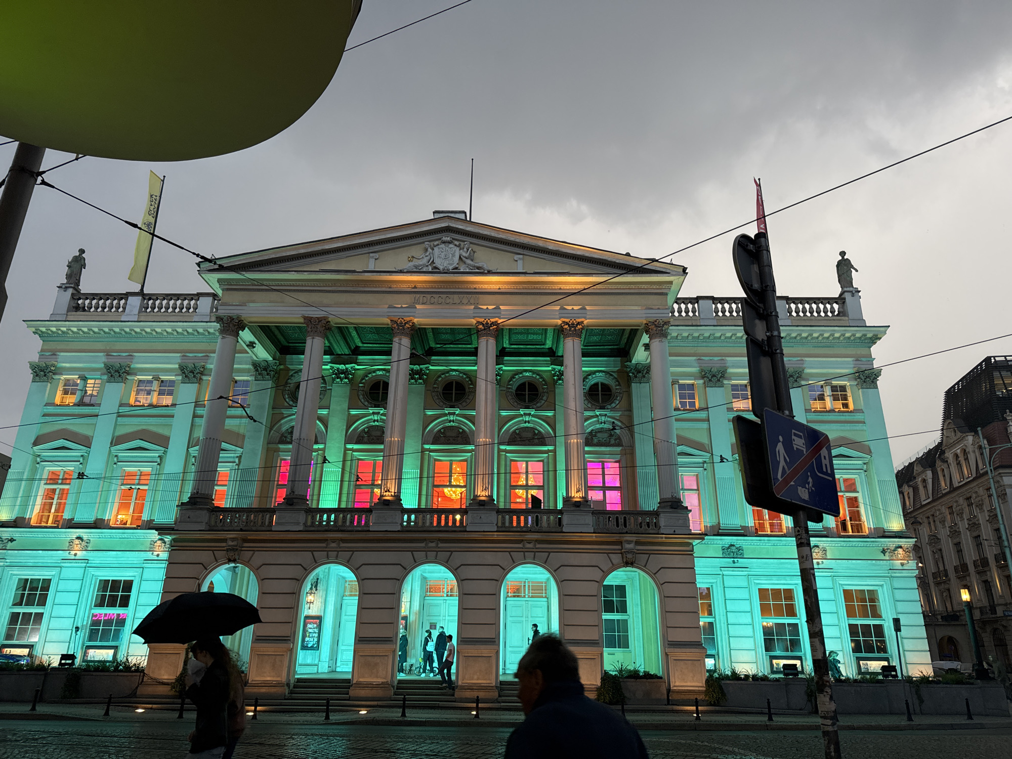 The image depicts the National Museum in Wrocław, Poland, illuminated with vibrant green and pink lights at dusk. The neoclassical building features a grand facade with columns, arches, and ornate architectural details. The lighting highlights the building's grandeur, creating a dramatic effect against the cloudy sky.
In the foreground, a person holding a black umbrella walks on the wet cobblestone street, suggesting recent or ongoing rain. The street is lined with posts and cables, indicating an urban setting with overhead tram lines. Further back, several individuals are visible near the entrance arches of the museum, some appearing to be entering or exiting the building.
A traffic sign indicating pedestrian and bus zones is prominently positioned on a pole in the middle right of the frame. The surrounding environment suggests a city square or main street, with other buildings visible in the background. The overall atmosphere is one of urban activity and cultural engagement, with the illuminated museum serving as the central focus.