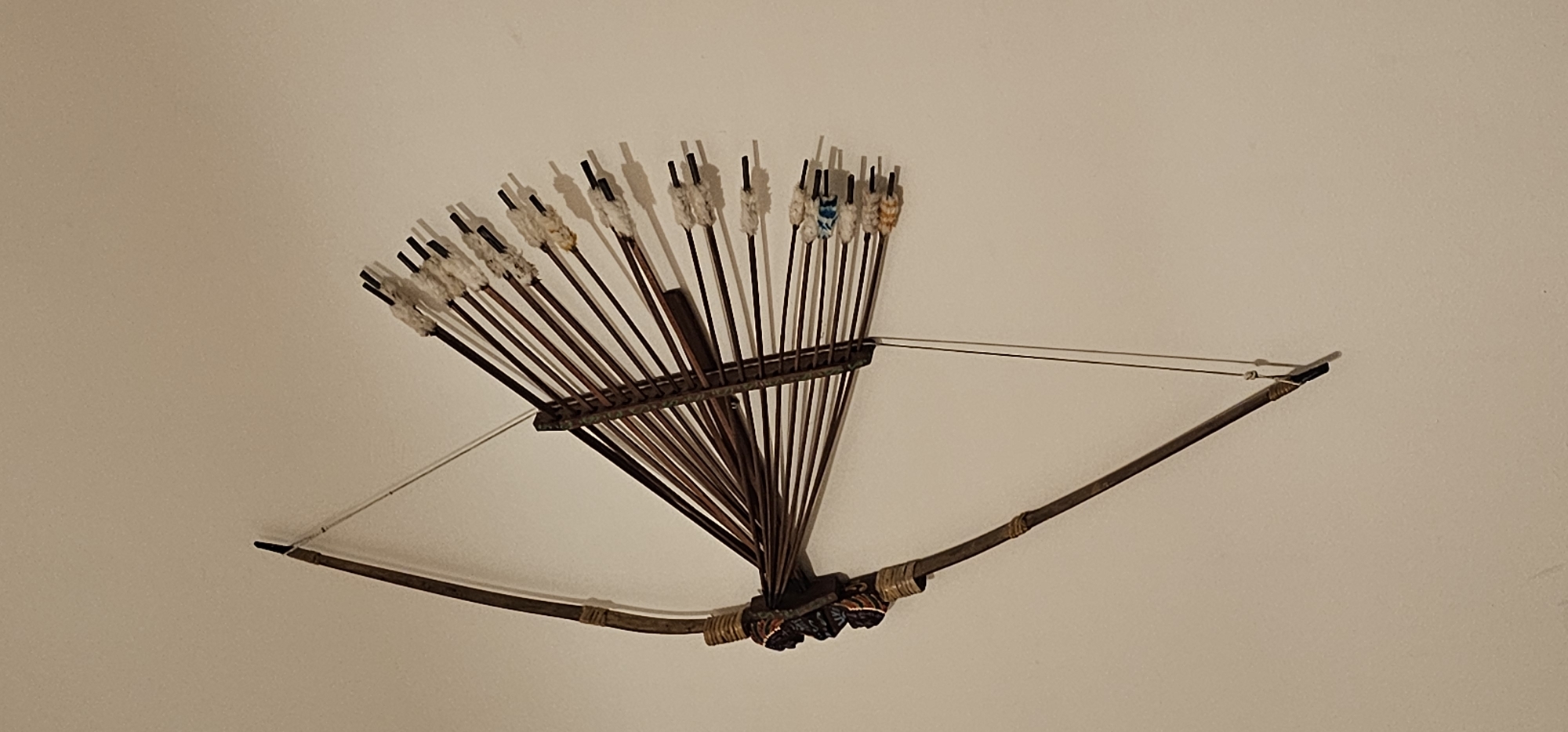 An ornate, antique bow and arrow set is mounted on a plain, light-colored wall. The bow, made of dark wood, is curved, with a string tautly stretched across it. At the center of the bow, where the arrows are fletched, is a dark, carved ornament that appears to be a human face. The arrows are fletched with white material, and some have hints of color, such as blue and orange, visible near the nock. The arrows are arranged in a fan-like pattern, held together by a darker wooden structure attached to the bow. The overall impression is of a decorative or historical item displayed as art. There is no indication of a specific location in Dogato, Italy, within the image itself.