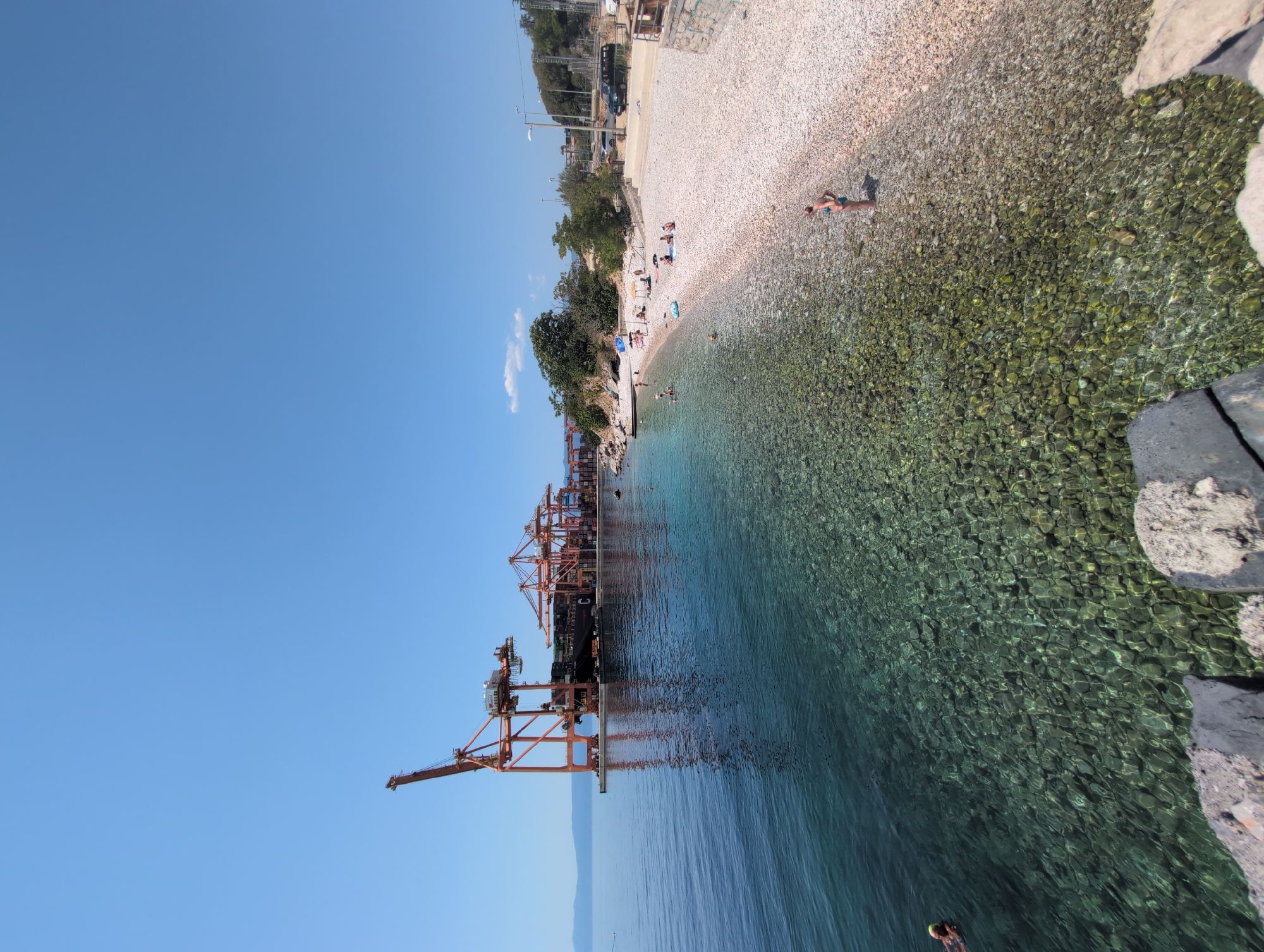 The image depicts a sunny day at a coastal area near the city of Rijeka, Croatia. In the background, a large cargo ship is docked at a port, with several tall, orange gantry cranes positioned over it, indicating a functioning industrial shipping facility. The water in the foreground is remarkably clear, allowing the rocky seabed to be visible. To the right of the water is a pebbled beach where several people are sunbathing and swimming. A few trees and vegetation are present on the shoreline, with a road and parked vehicles visible further inland. The sky is a clear, bright blue.