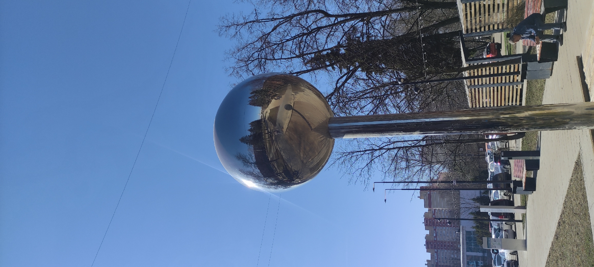 A street scene in Fryazino, Russia features a large, shiny, spherical sculpture mounted on a cylindrical base. The sculpture reflects the surrounding environment including trees, buildings, and the clear blue sky. The street has benches and lampposts, with parked cars along the side. Buildings of moderate height, constructed with red brick and concrete, are visible in the background, typical of urban residential areas. One person is visible interacting with one of the benches. The trees are bare of leaves, suggesting late winter or early spring.