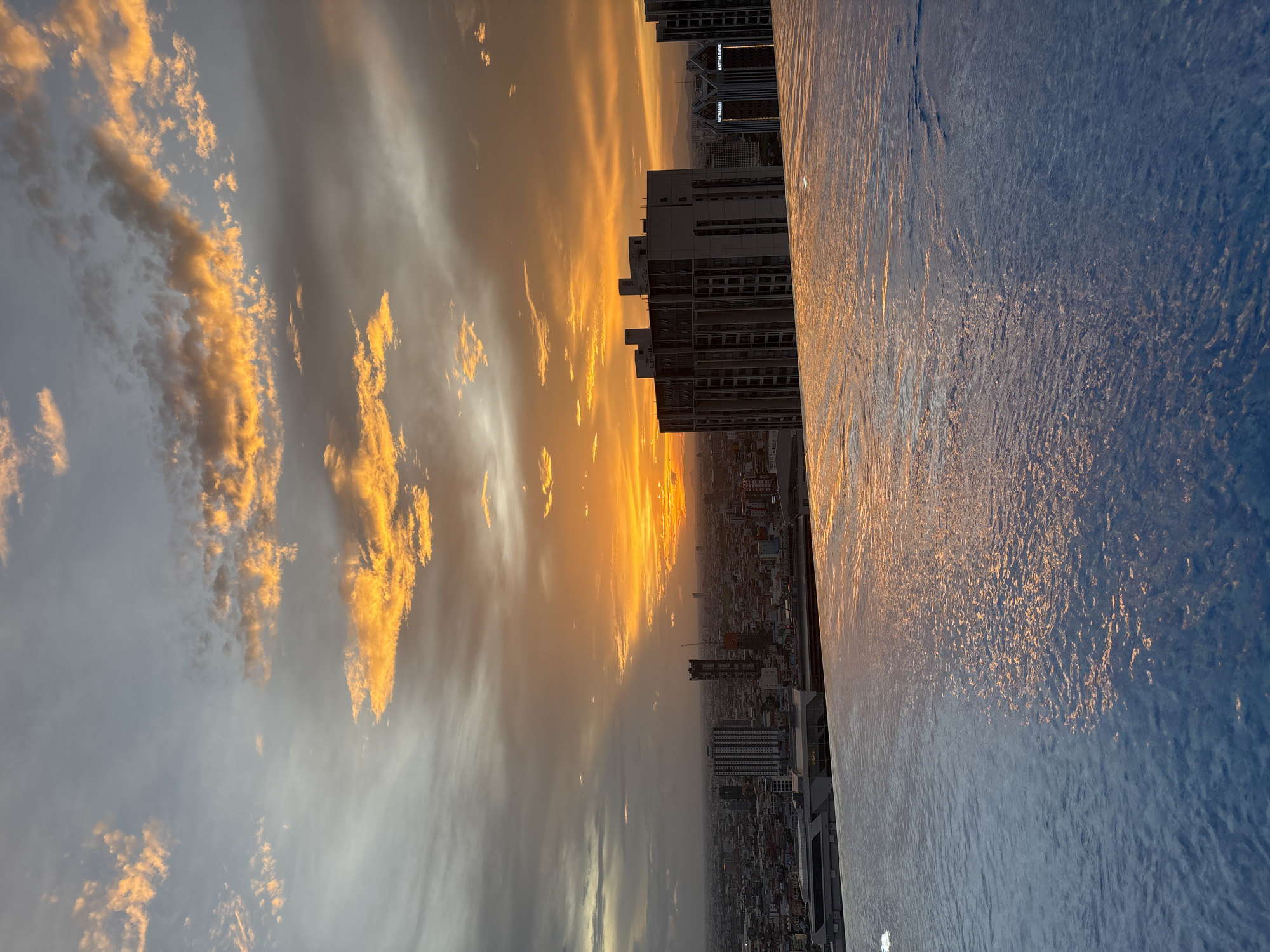 The image shows an infinity pool overlooking a city skyline at sunset. The sky is filled with dramatic clouds in shades of orange, yellow, and gray, illuminated by the setting sun. The city below is a mix of buildings, including high-rise apartments and office buildings. The pool's surface reflects the colors of the sky, creating a shimmering effect. The location is identified as the city of Khan Mean Chey, Cambodia, likely from a rooftop vantage point. There are no people visible in the image. The overall mood is serene and awe-inspiring, capturing the beauty of a sunset over an urban landscape.