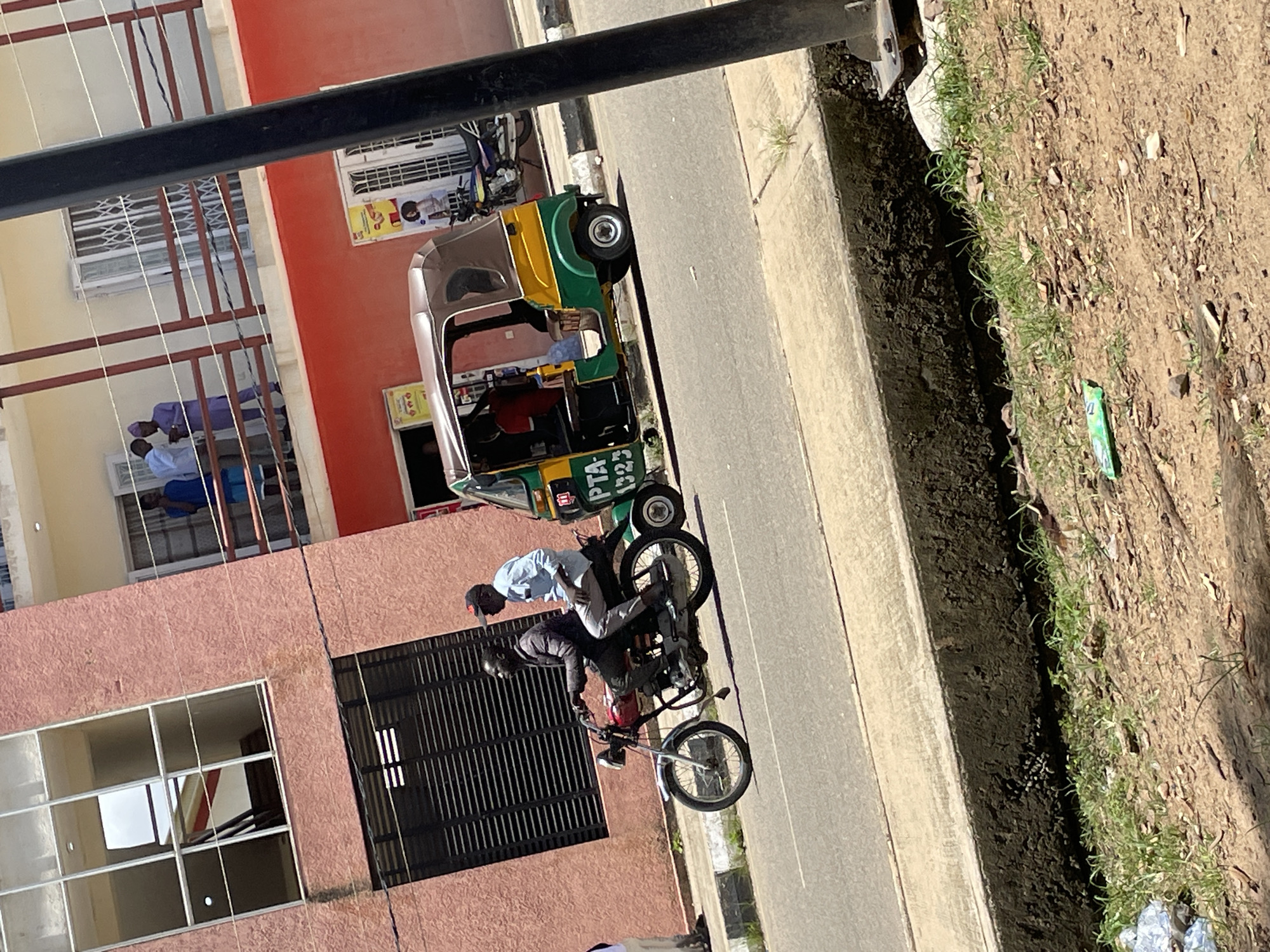 In Jos, Nigeria, a motorcycle with two male occupants, a driver and a passenger, travels on an asphalt road. The driver, wearing a dark jacket and black pants, operates the red motorcycle. The passenger, in a light blue shirt and gray pants, sits behind the driver. A green and yellow auto-rickshaw follows closely behind the motorcycle. The scene unfolds in front of a multi-story building with a pink facade and large glass windows. On an upper level balcony, several individuals are visible. The environment includes a concrete sidewalk, a drainage ditch, and a dirt area with sparse vegetation.