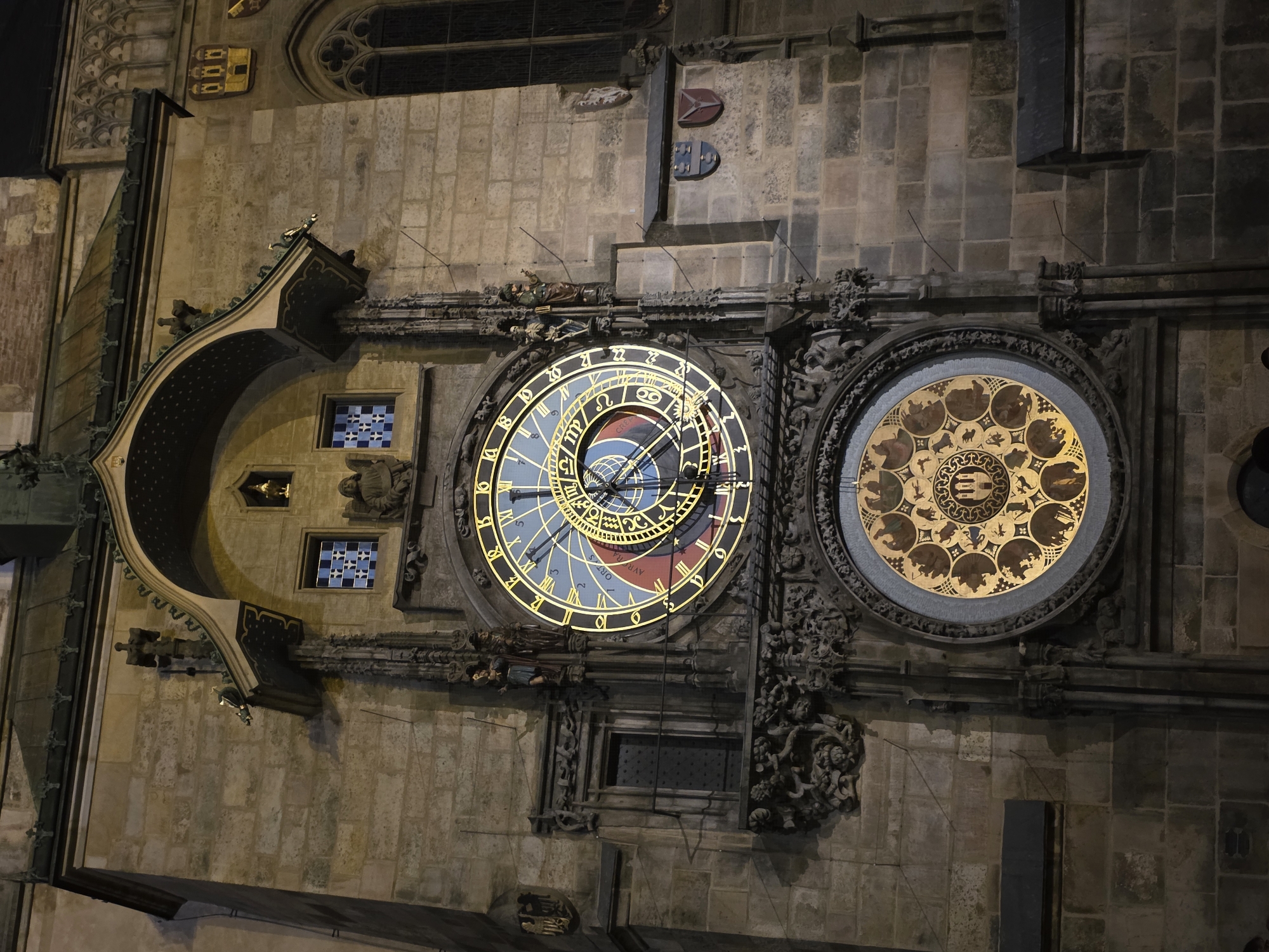 The image shows a close-up, low-angle shot of the Prague Astronomical Clock tower at night. The clock face, with its Roman numerals and astronomical dial, is illuminated and prominently featured. Below it, a second, smaller dial with zodiac symbols is also visible. The clock tower is constructed of stone blocks and features intricate architectural details, including decorative carvings, statues, and a small arched window with blue and white stained glass. Gargoyles and ornate metalwork are also visible on the building. The overall lighting suggests the image was taken after dusk, with artificial lights illuminating the clock and parts of the facade.