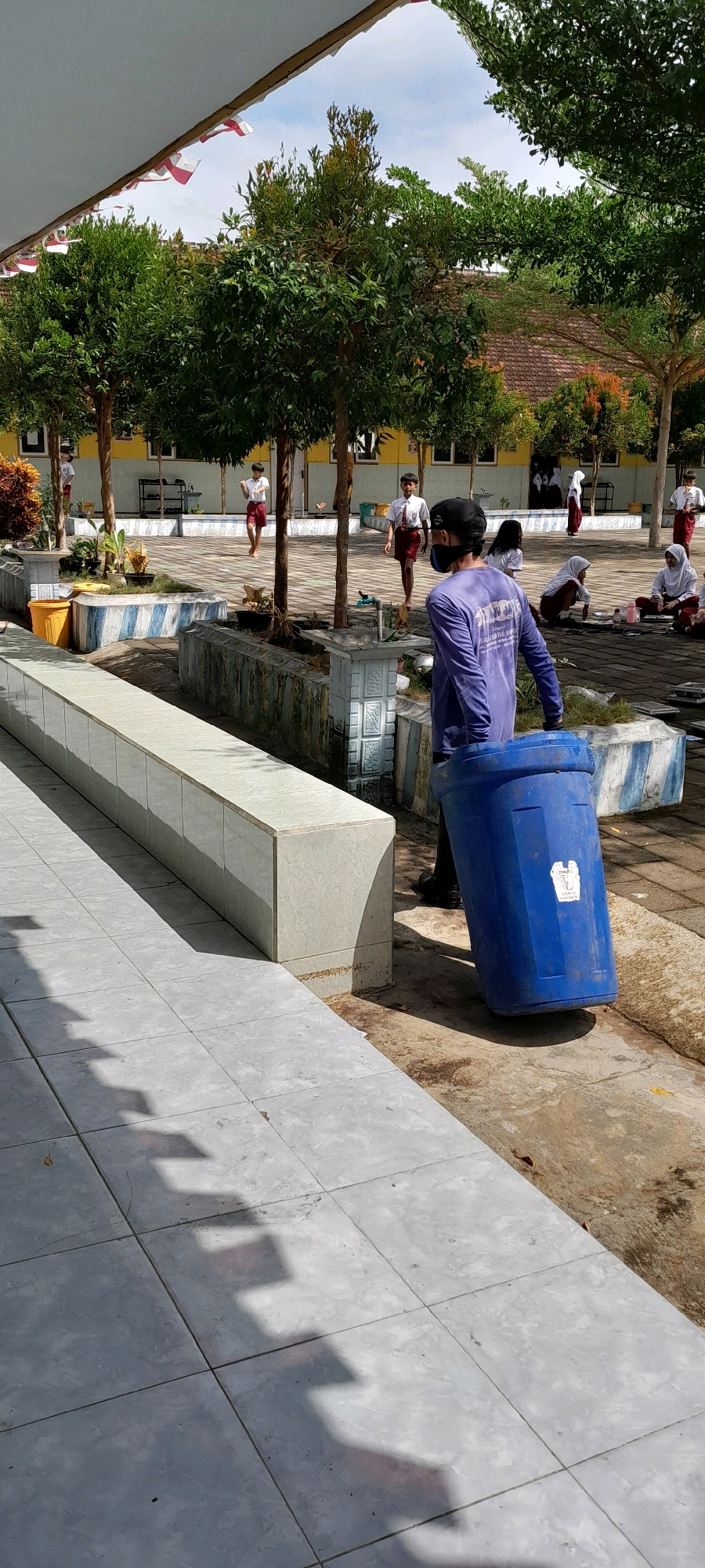An outdoor, daytime scene depicts a person in the foreground wearing a purple long-sleeved shirt, a black face mask, and a black baseball cap, pushing a large blue trash bin. The bin is tilted on its side, being rolled on its base. The person's back is to the camera. Behind this person, several school-aged children in white shirts and red shorts are visible. Some are walking, while others are sitting on the ground, seemingly engaged in an activity. The environment appears to be a schoolyard, with trees, potted plants, and a building with yellow walls in the background. Red and white Indonesian flags are strung across a white overhang in the upper left of the frame. The ground is paved with grey tiles. This scene is located in Rogojampi, Indonesia, as indicated by the context of the image.