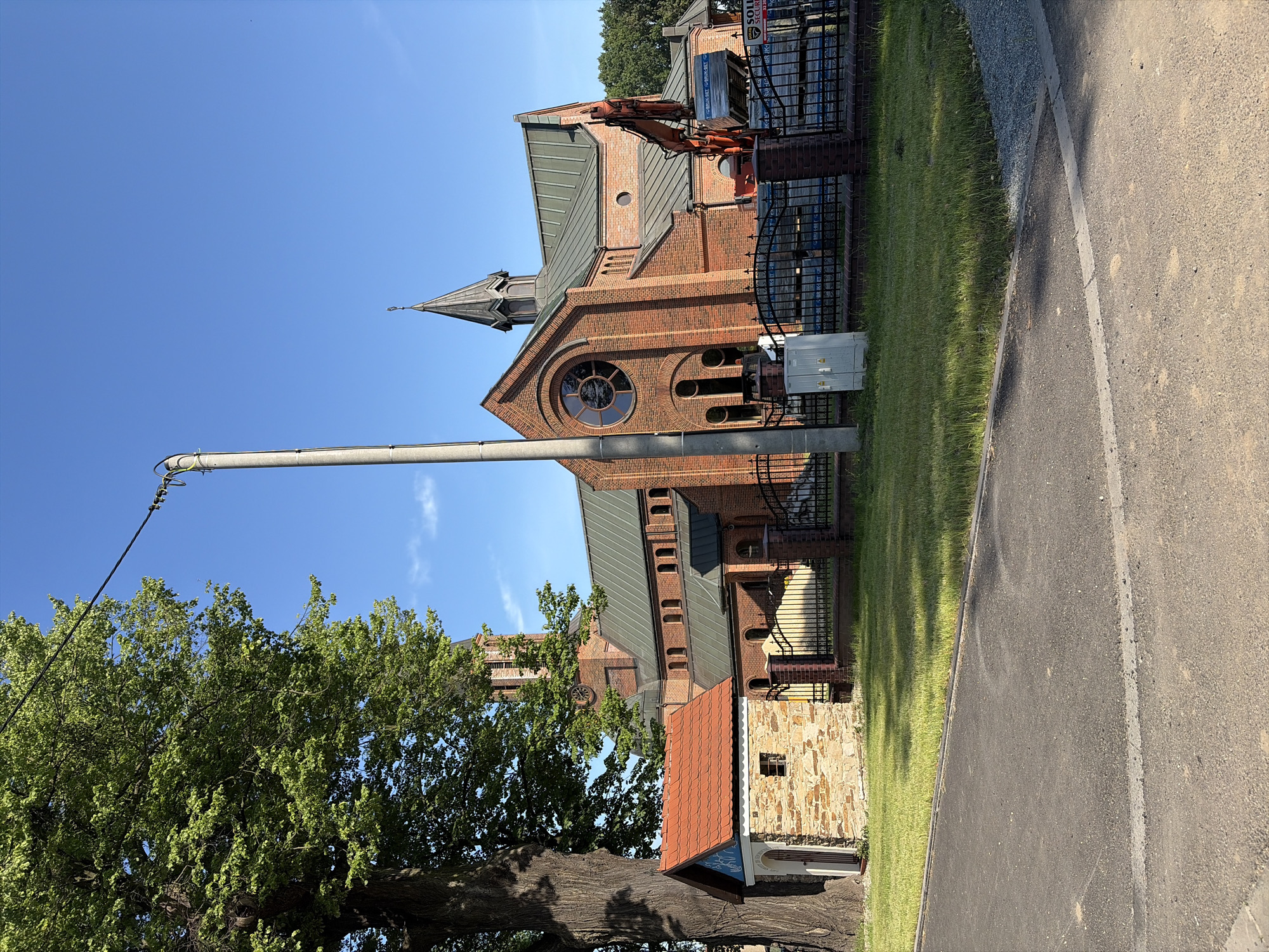 The image depicts the Church of St. Hedwig in Stanowice, Poland, under what appears to be ongoing construction or maintenance. The brick church features a prominent steeple and a large circular stained-glass window above the main entrance. An orange excavator is positioned in front of the church, its bucket raised, suggesting recent activity. Several stacks of building materials, possibly bricks or pavers, are visible behind a black metal fence surrounding the church grounds. To the left of the church, a small, rustic stone structure with a red-tiled roof stands near a large, mature tree. The scene is illuminated by bright sunlight on a clear blue sky. A concrete utility pole with electrical wires runs vertically through the left side of the frame. The area in the foreground shows an asphalt road with a light-colored stripe.